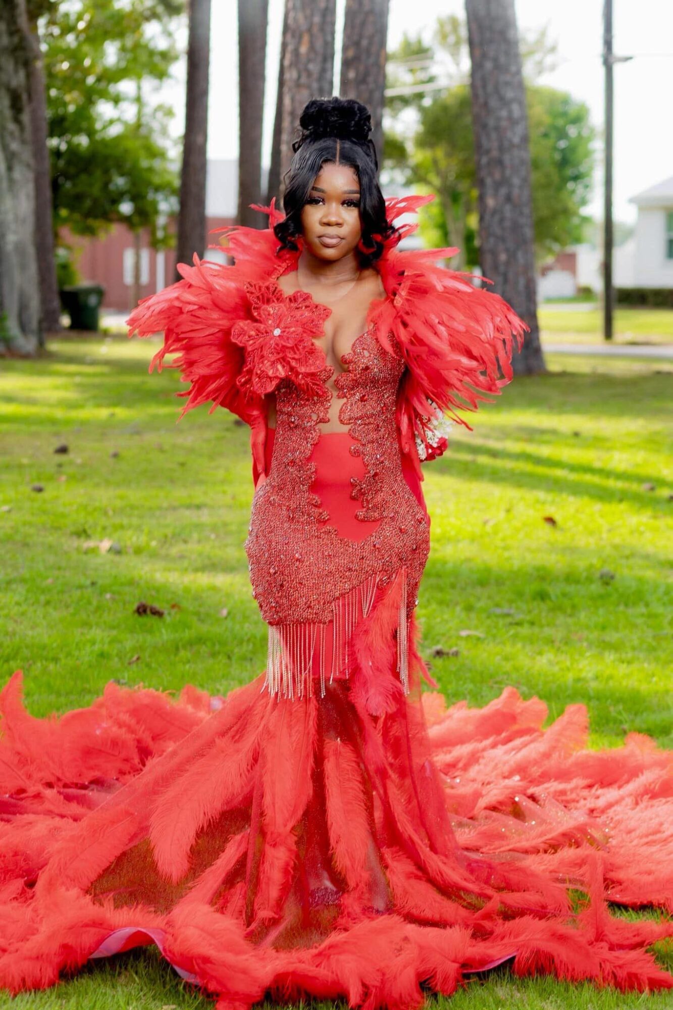 Woman in a red gown with feathered shoulders standing outdoors on grass with trees and houses in background.