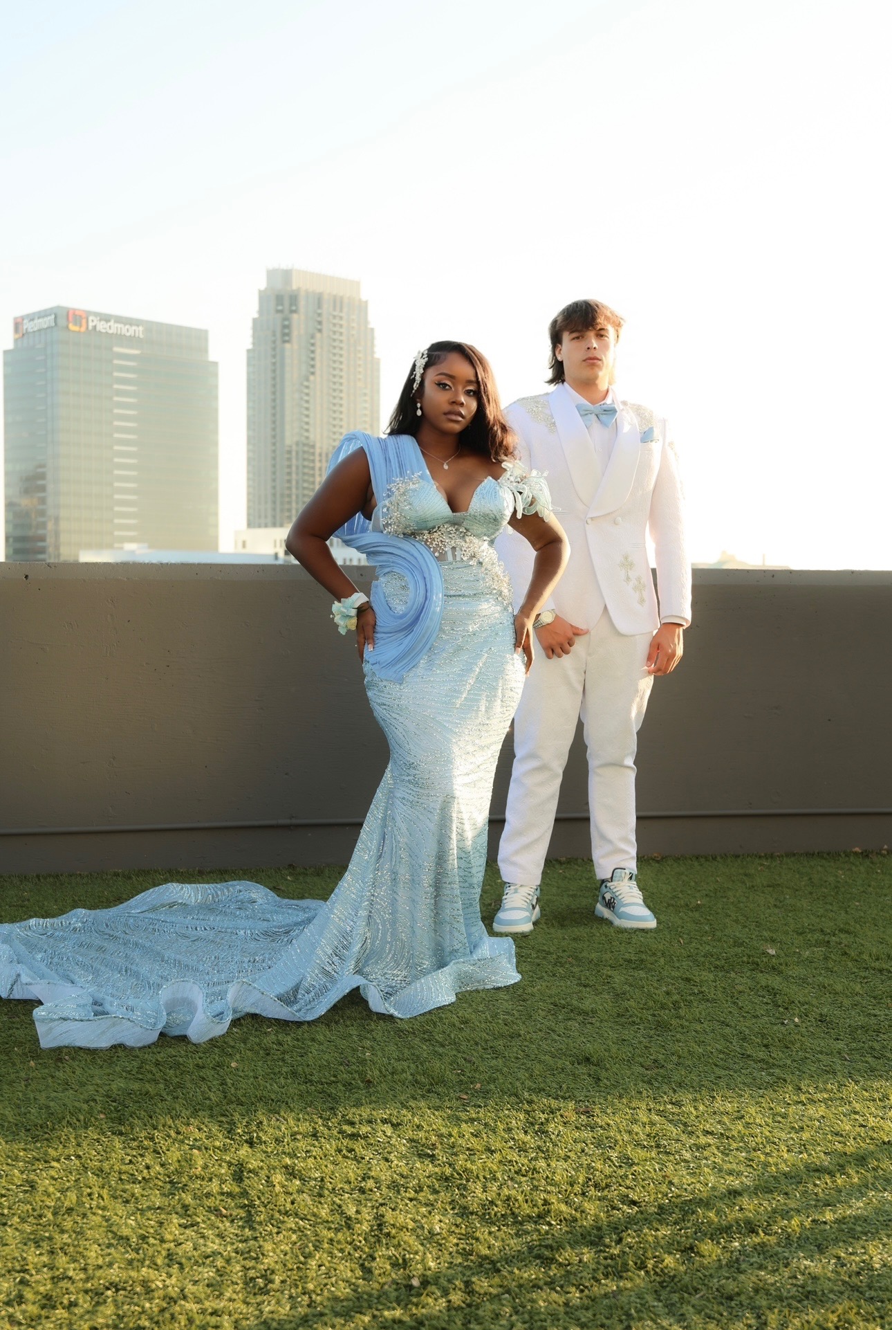 A woman in a light blue gown and a man in a white suit stand outdoors on grass with city buildings in the background.