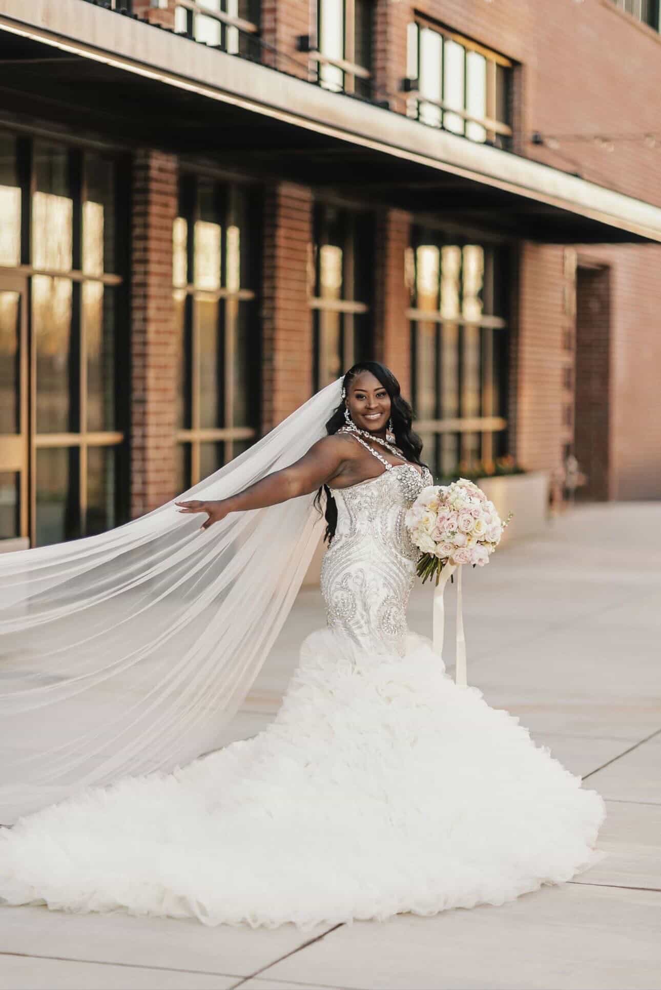 Bride in a white wedding gown holding a bouquet, standing outdoors with a building in the background.