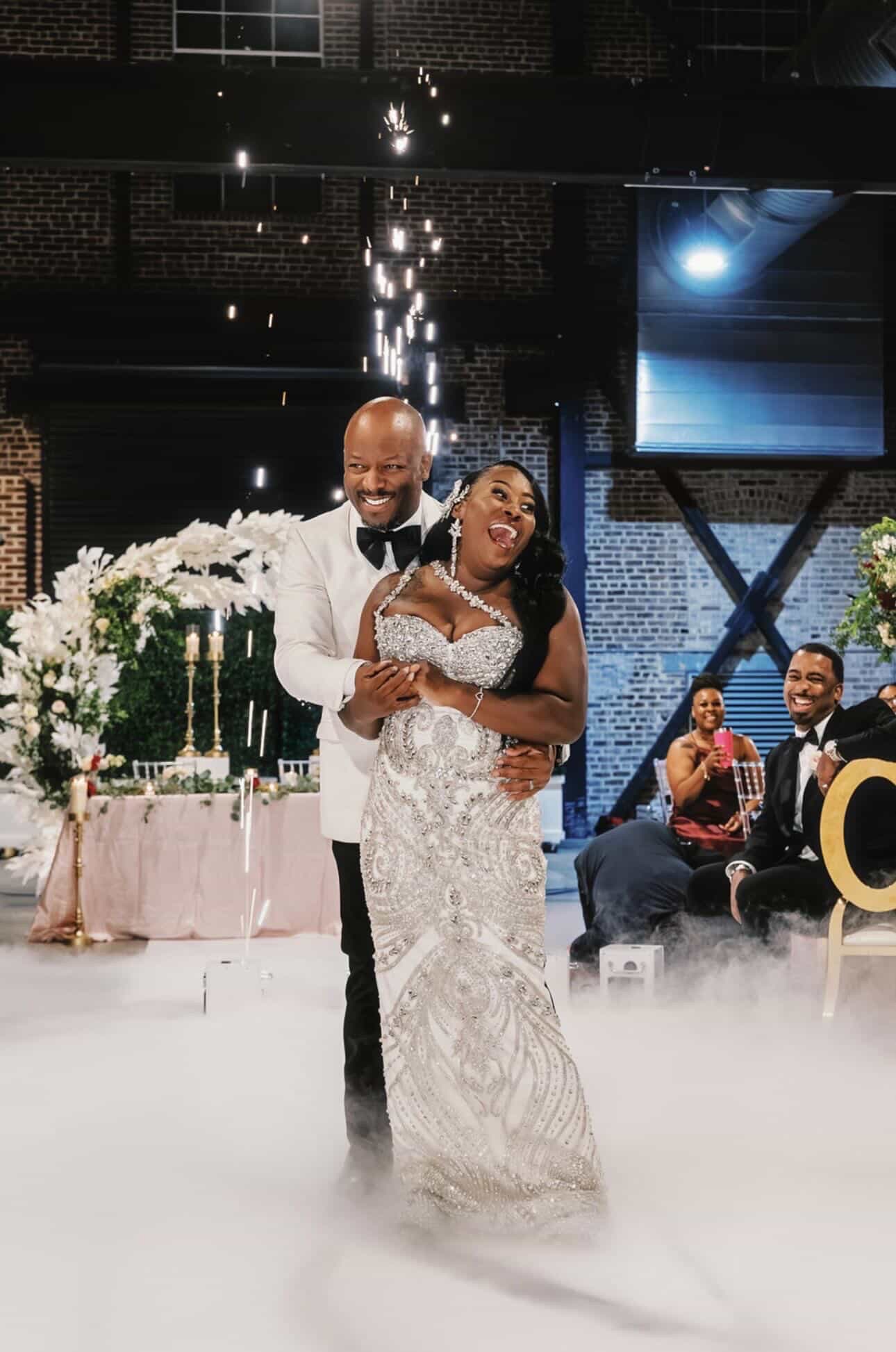 Couple dancing at wedding reception, smiling, with guests and decorations in background.