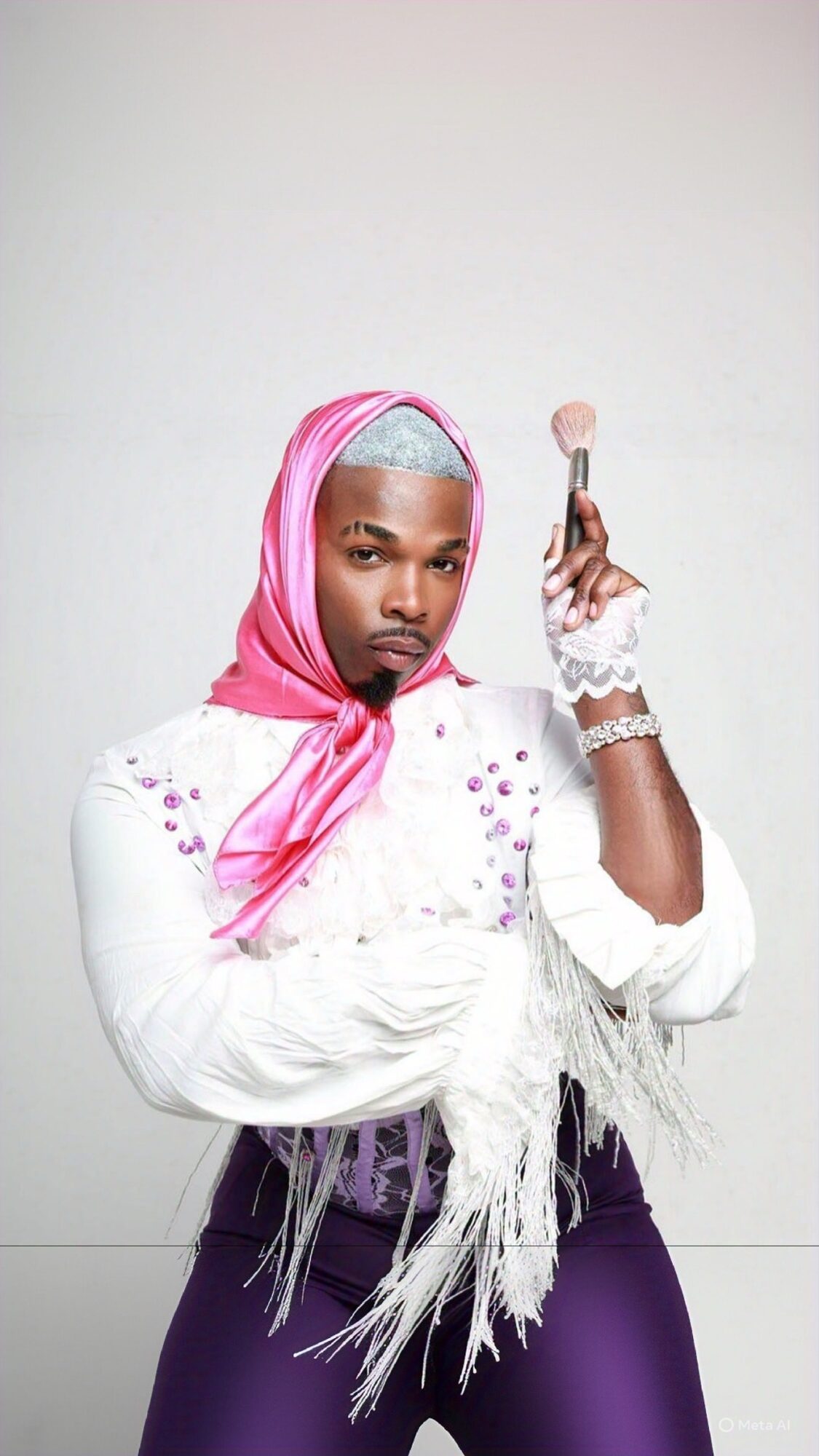 Person with pink and silver headscarf holding a makeup brush, wearing a white fringed top and purple pants, standing against a plain background.