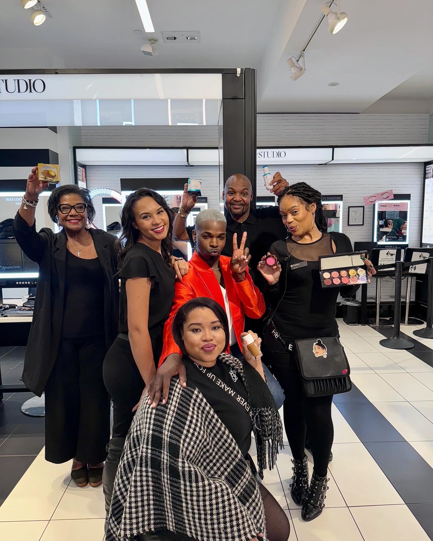 Group of six people inside a store, smiling and posing for the photo, some making peace signs, with makeup products in the background.