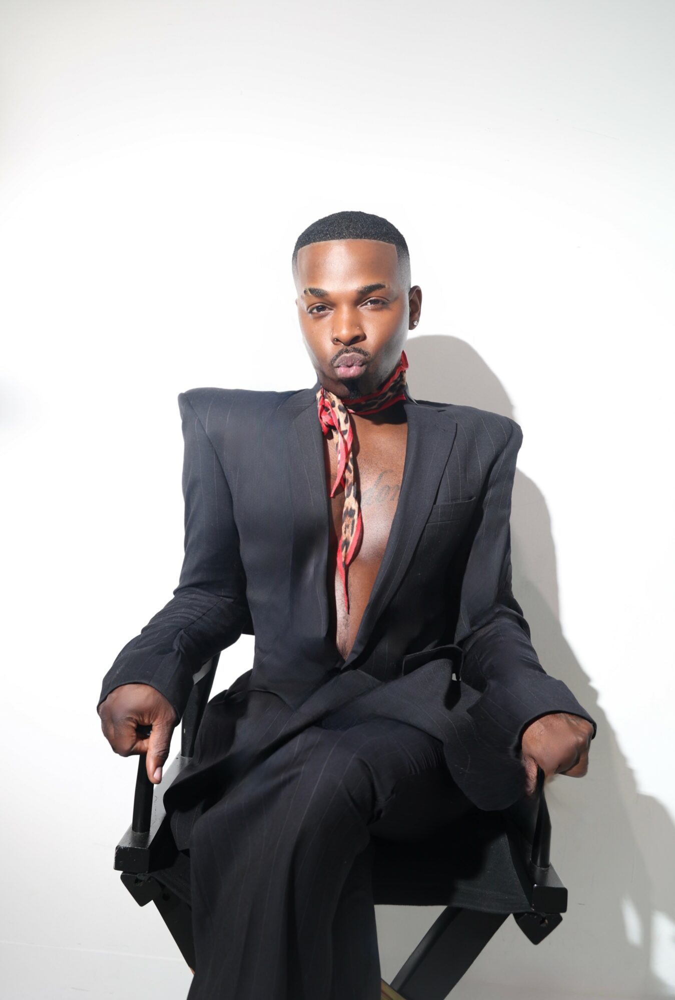 Person with short hair and beard wearing a black blazer and pants, sitting on a chair against a white background.