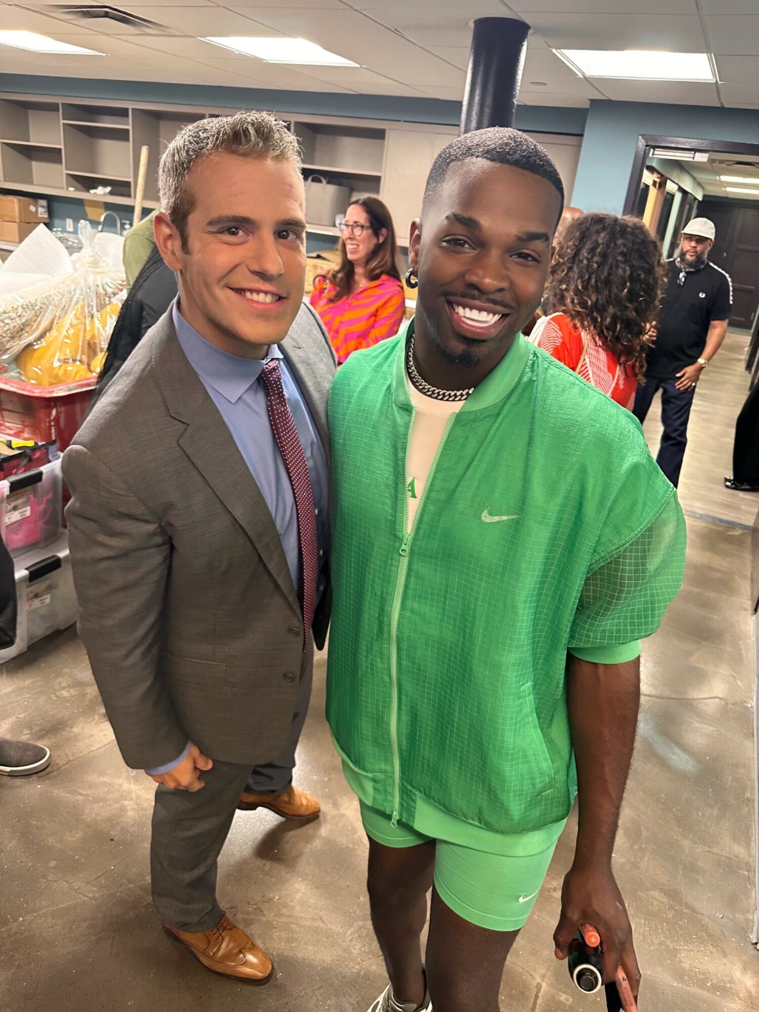 Two men smiling indoors, one in a suit and the other in a green jacket and shorts, with people in the background.