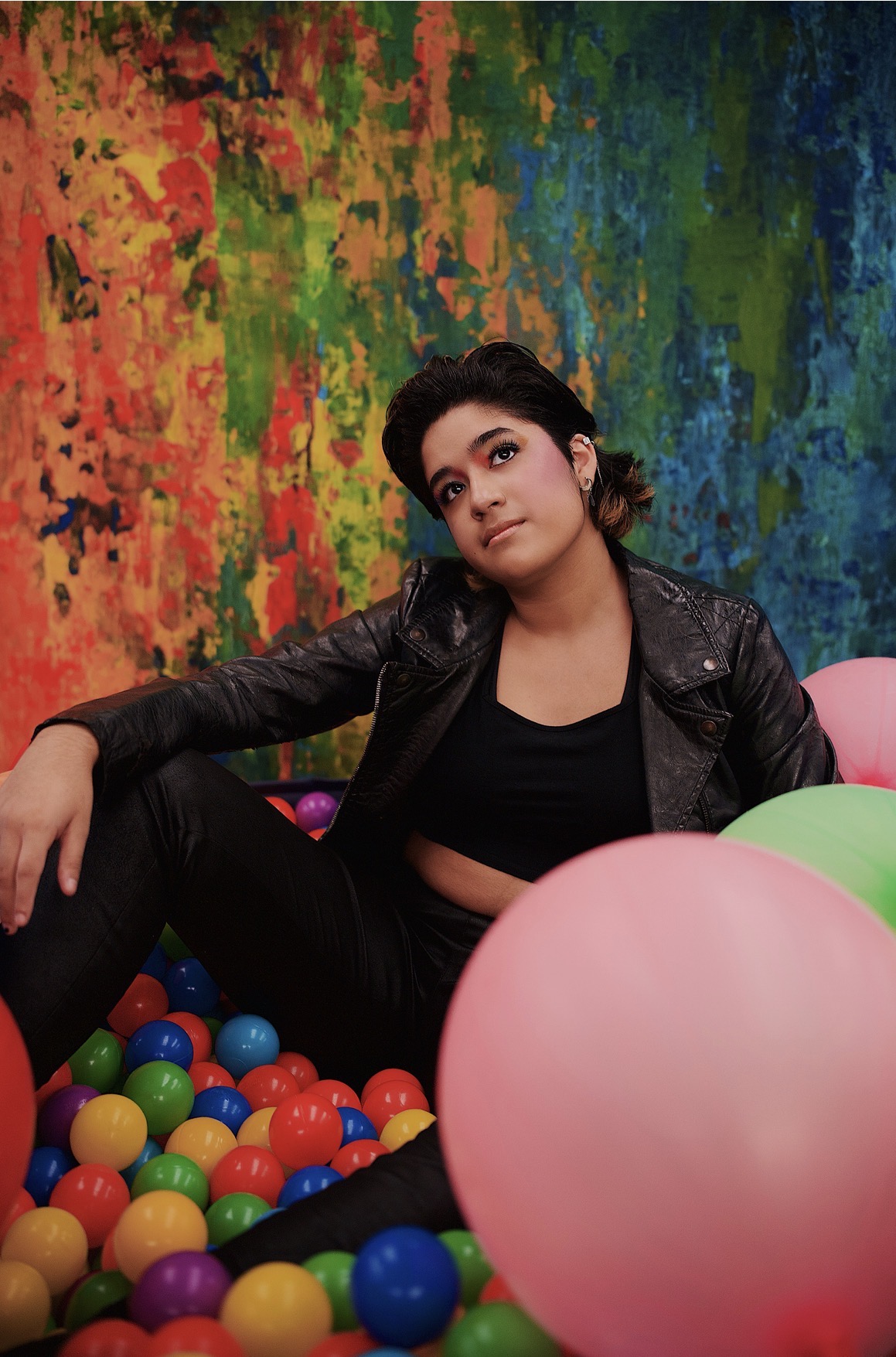 Young woman with dark hair in a black leather jacket sitting among colorful balloons against a vibrant, multicolored background.