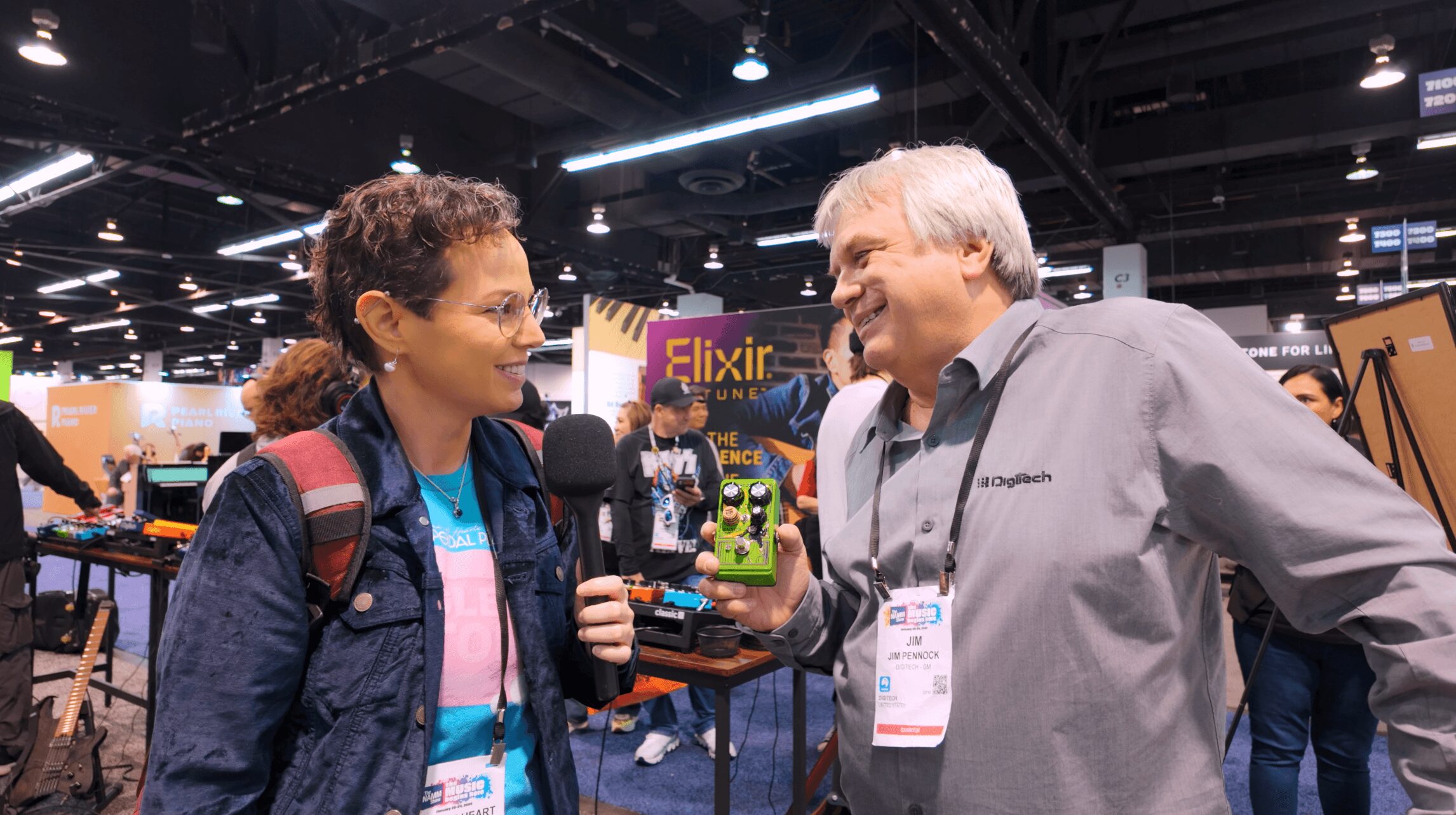 Two people talking at a convention, one holding a small green object, with booths and signs in the background.