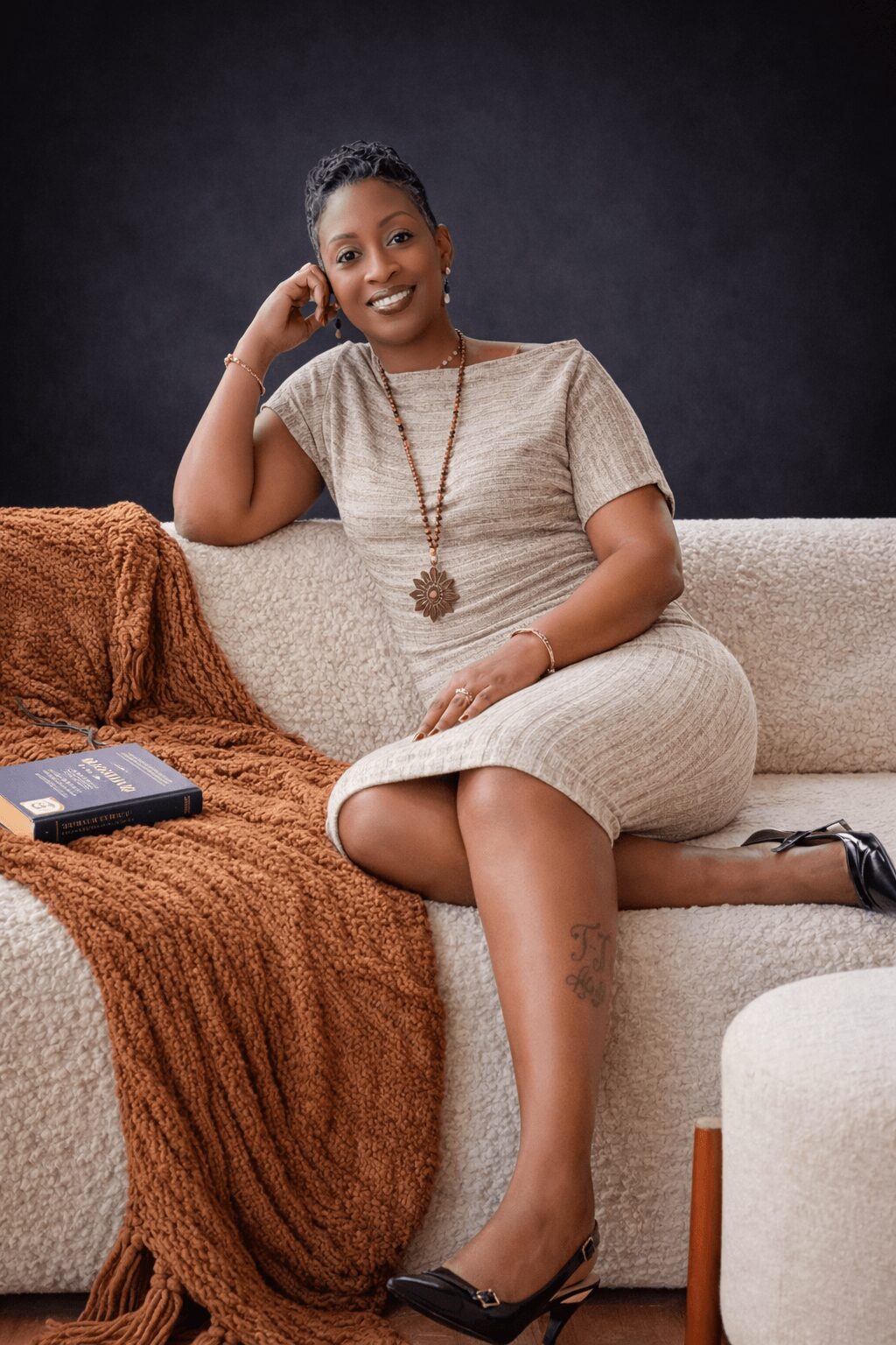 Woman sitting on sofa, smiling, wearing a light-colored dress, with a book and blanket nearby.