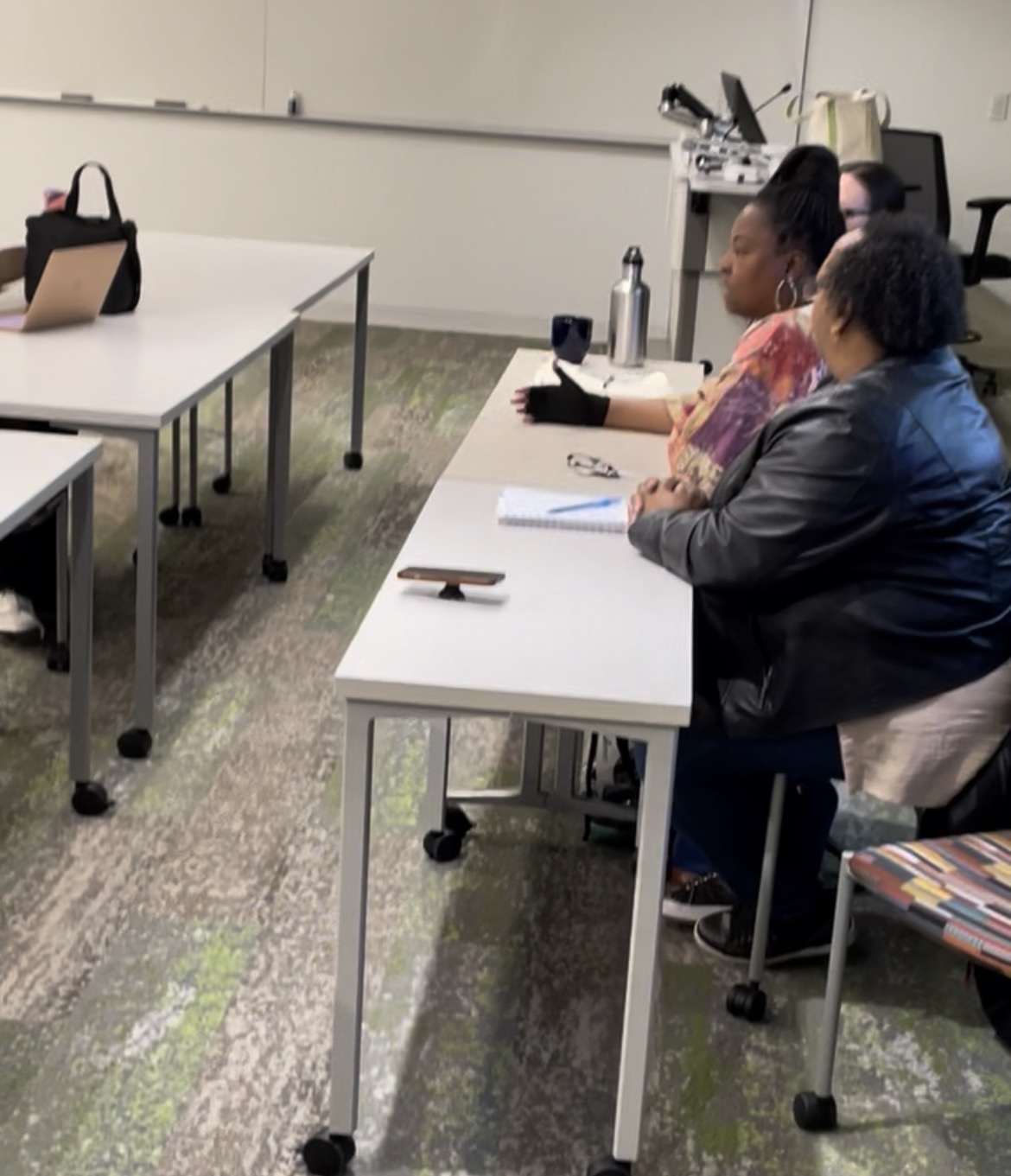 Two women sit at a white table in a classroom or meeting room, with notebooks and a water bottle, facing forward.
