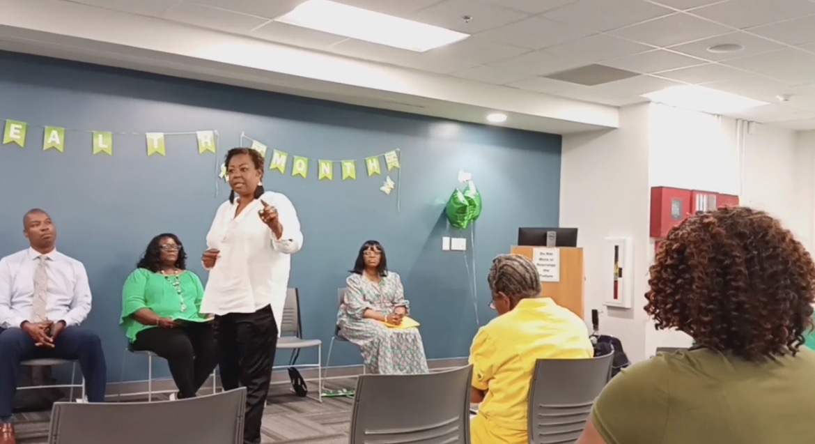 A woman stands and speaks to a seated group in a room with blue walls and decorations, including a green balloon and yellow banners.
