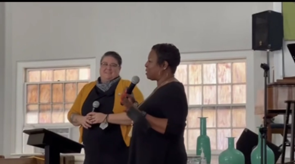 Two women speaking into a microphone in a room with large windows and decorative vases.