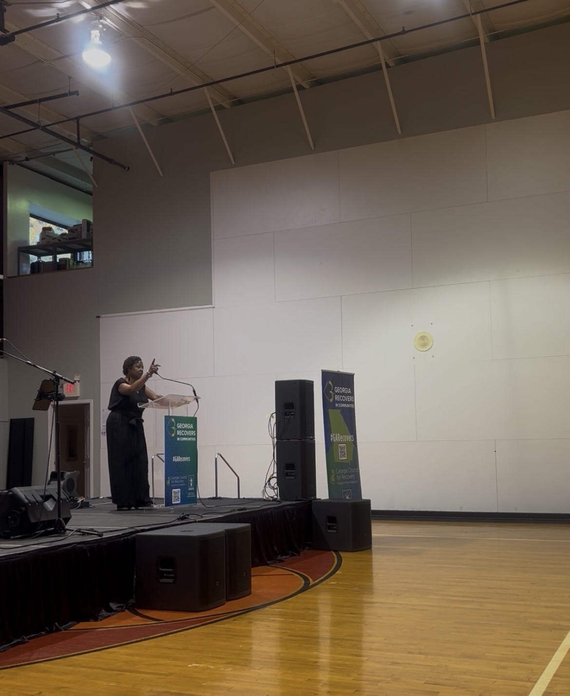Person playing violin on stage with speakers and banners, empty wall background, wooden floor, ceiling lights.