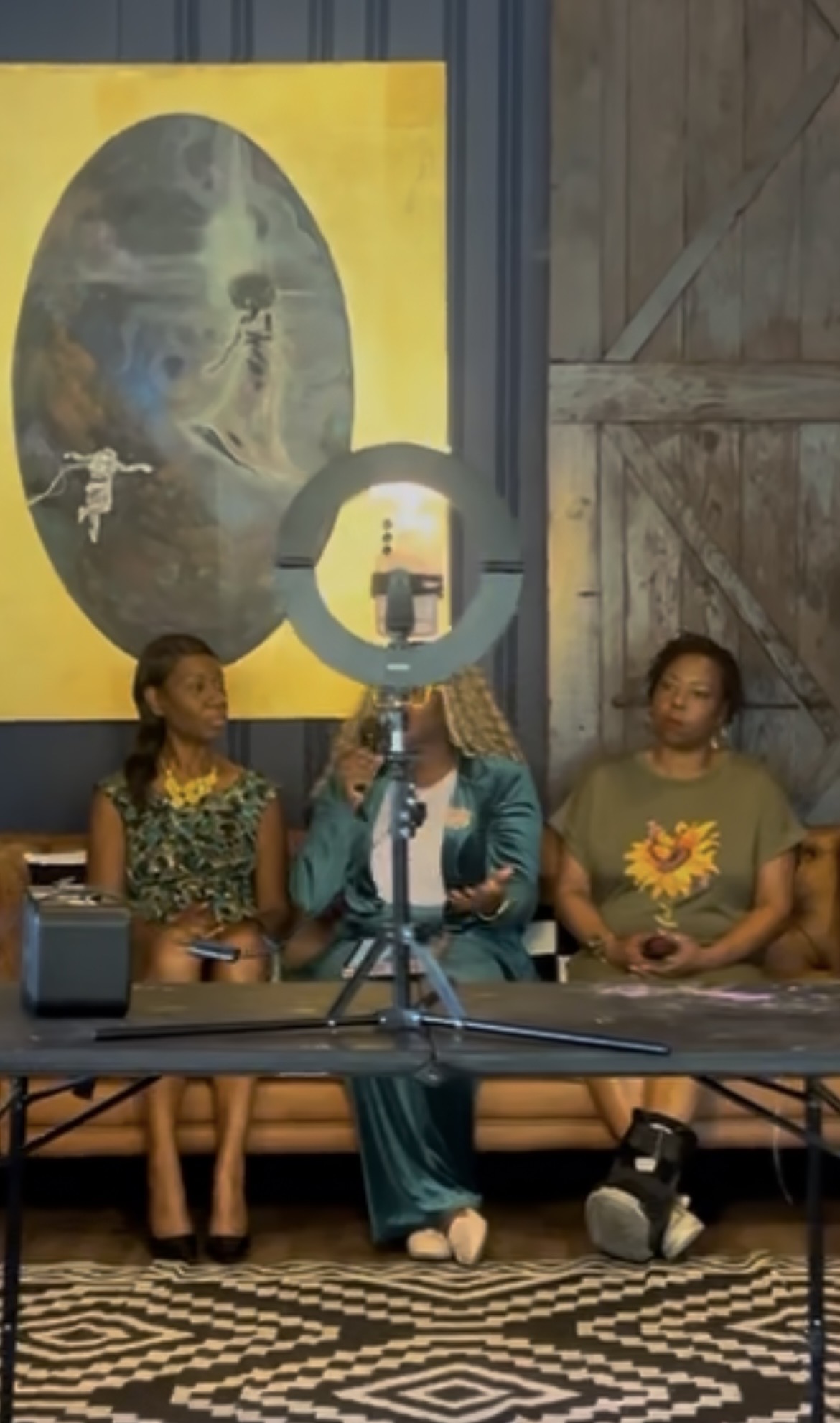 Three women sit at a table with a ring light, in front of a large painting on a wooden wall.