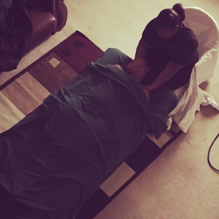Two people sitting on a bed in a room, one facing away and the other facing forward, with a colorful rug nearby.