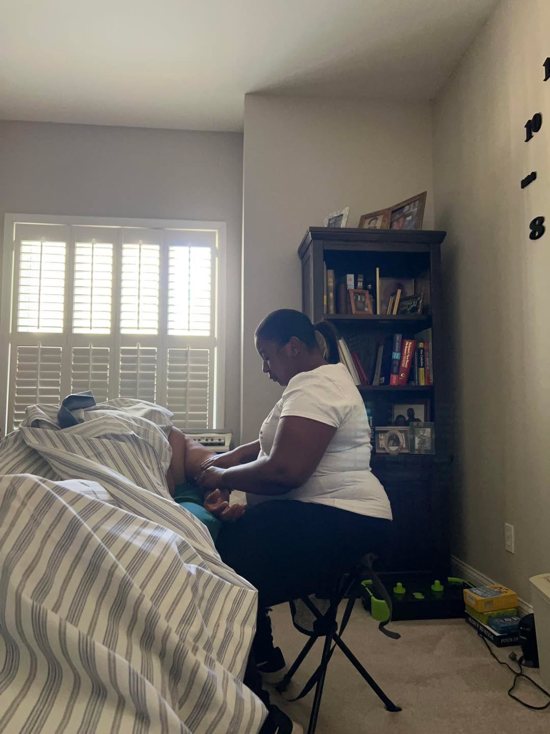 Person sitting on a chair next to a bed, reading a book in a room with a window and bookshelf.