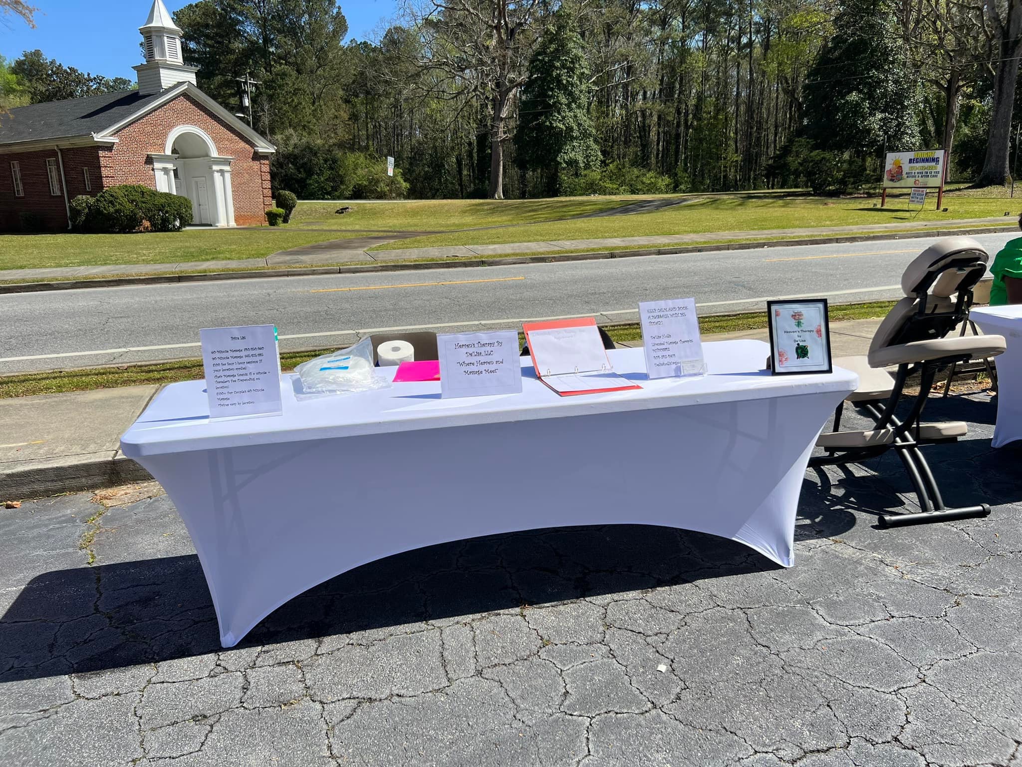 Outdoor table with informational signs, a tablet, and a stroller on pavement, with a church and trees in background.