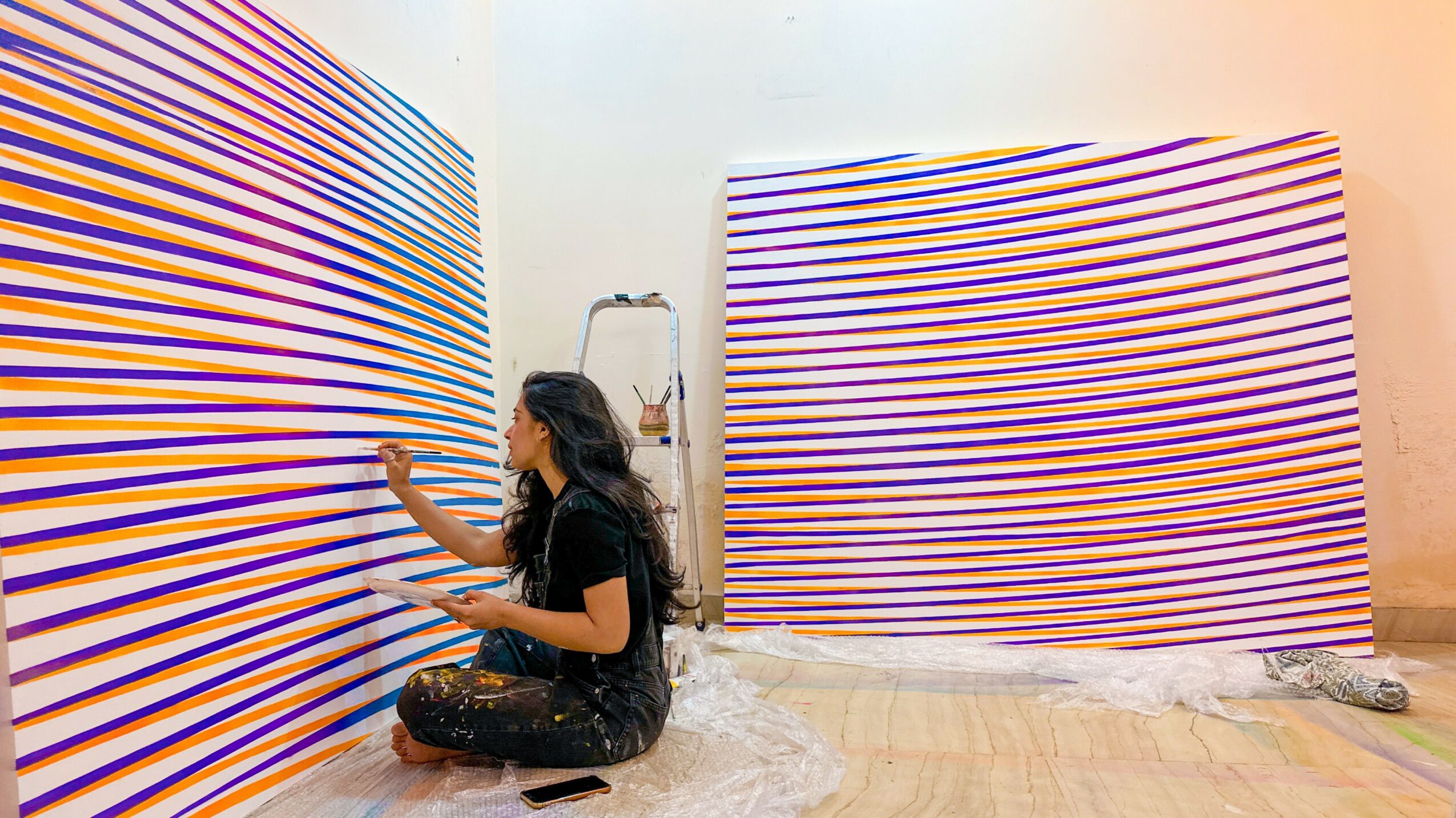 Person sitting on floor painting colorful horizontal stripes on large curved walls in an art studio.