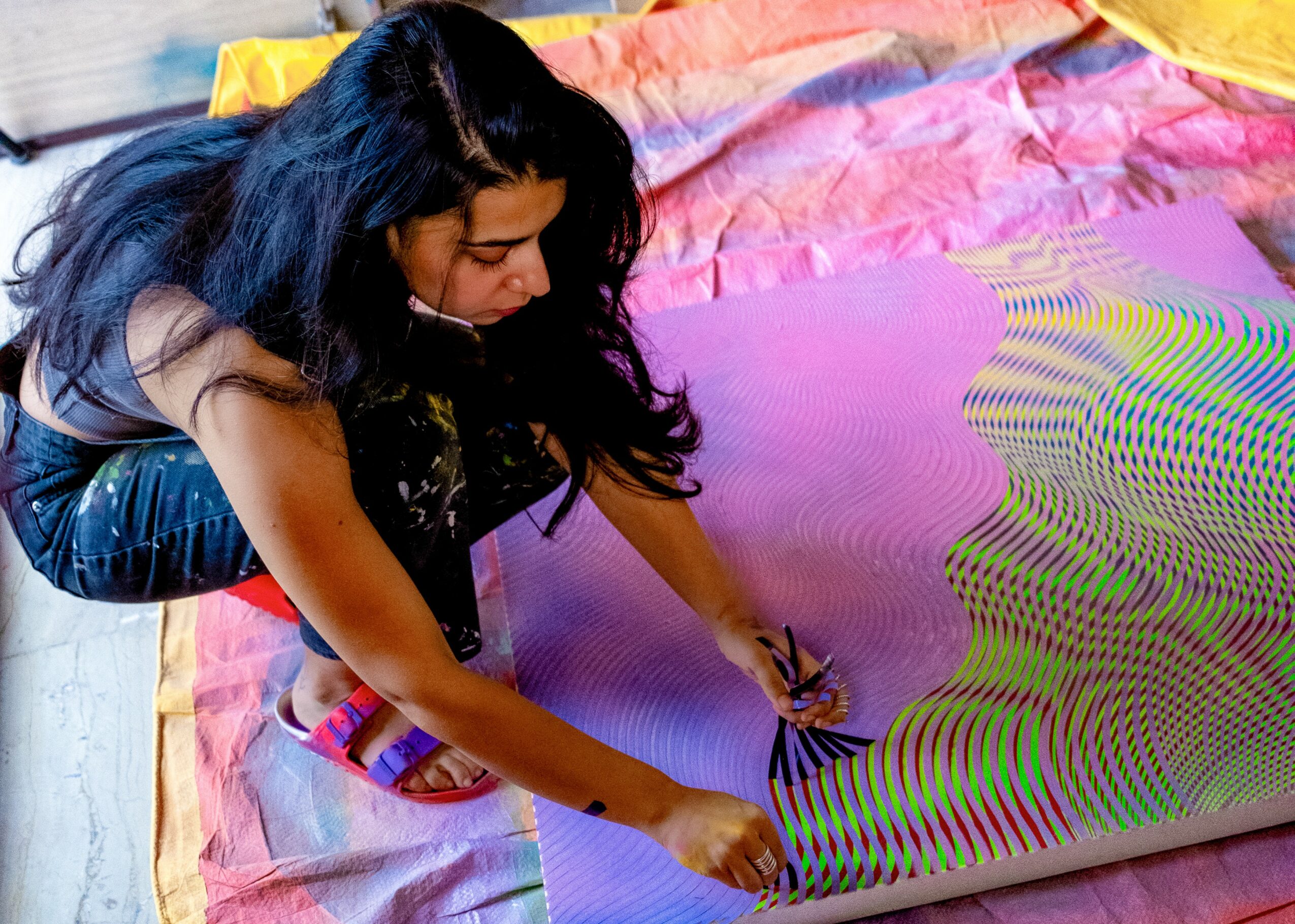 Person with long dark hair working on colorful, patterned artwork on a large sheet of paper.