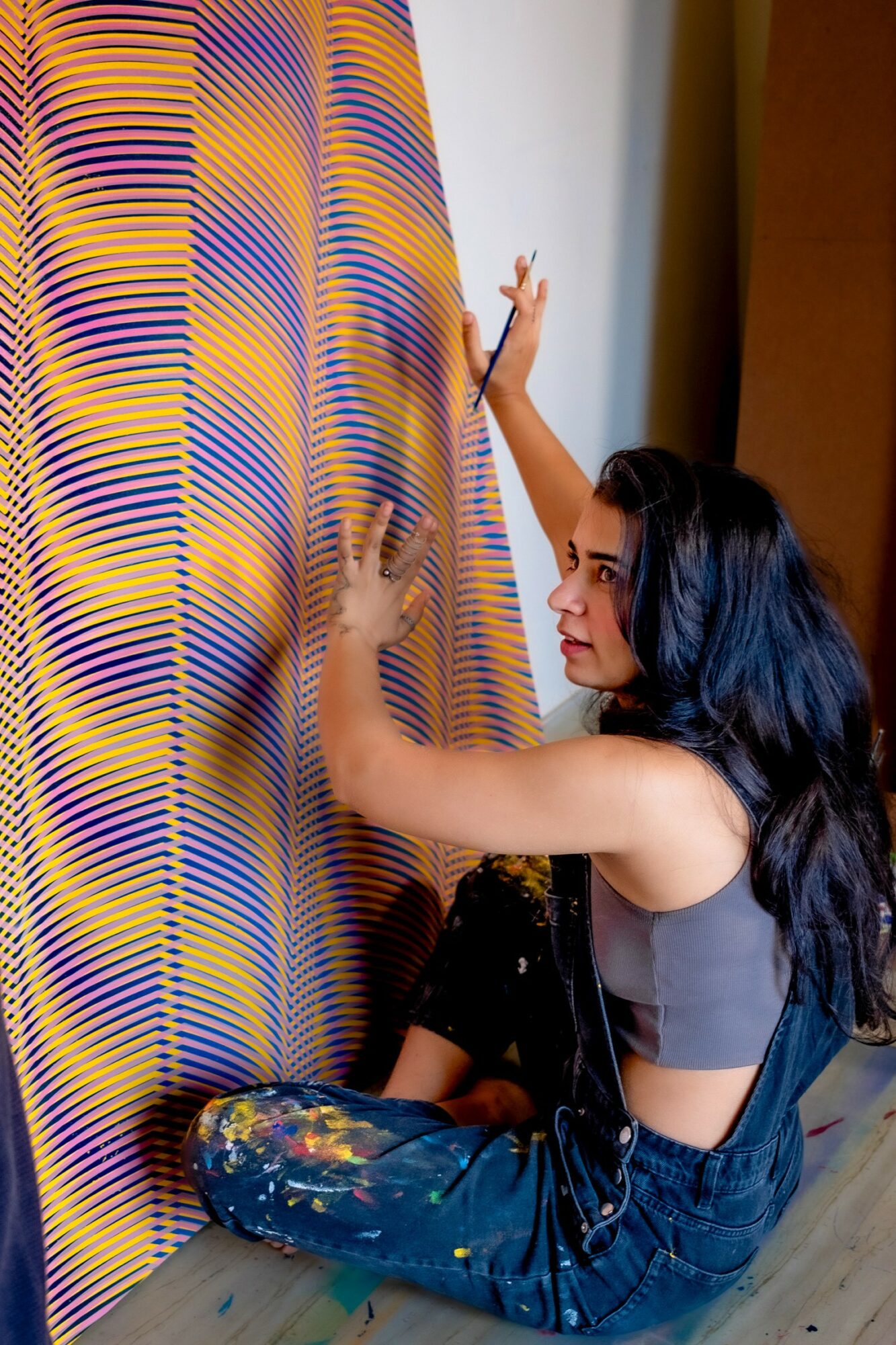 Young woman with long dark hair reaching up to touch a colorful, striped curtain, sitting on the floor.