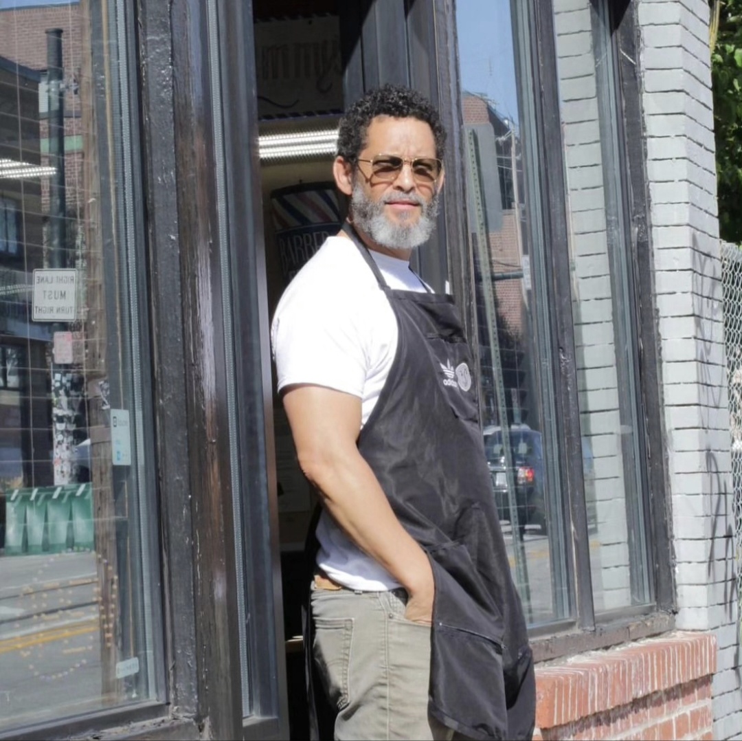 Man with glasses and beard standing outside a shop, smiling, wearing a white t-shirt and black apron, hands in pockets.