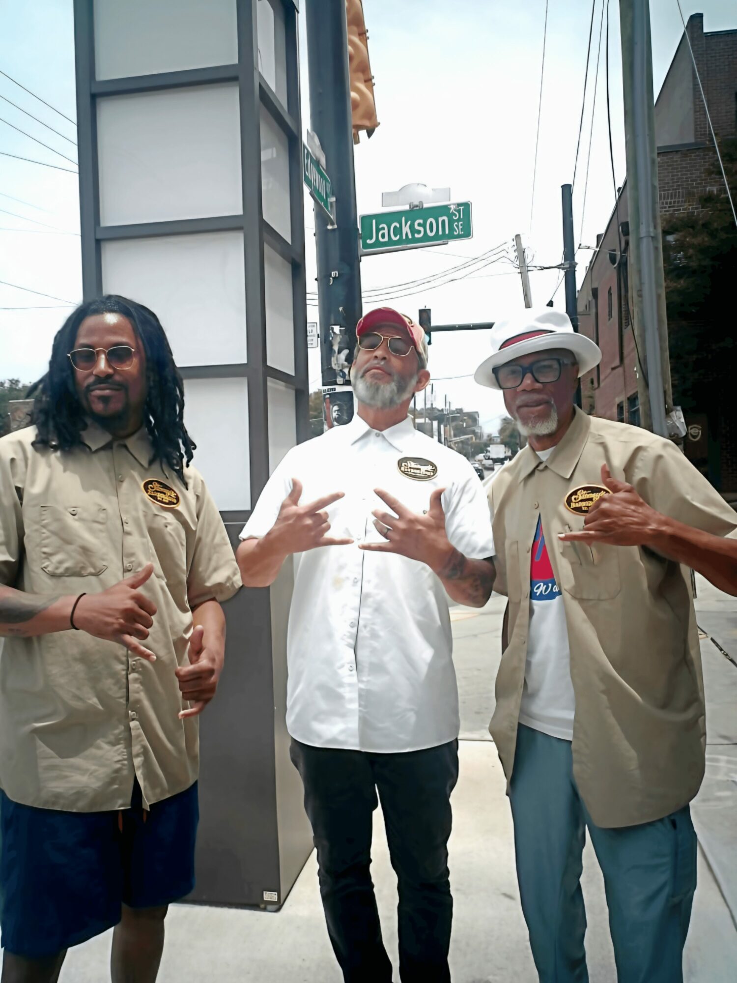 Three men standing outdoors near a street sign that reads 'Jackson'. They are wearing casual shirts and hats, posing for the photo.