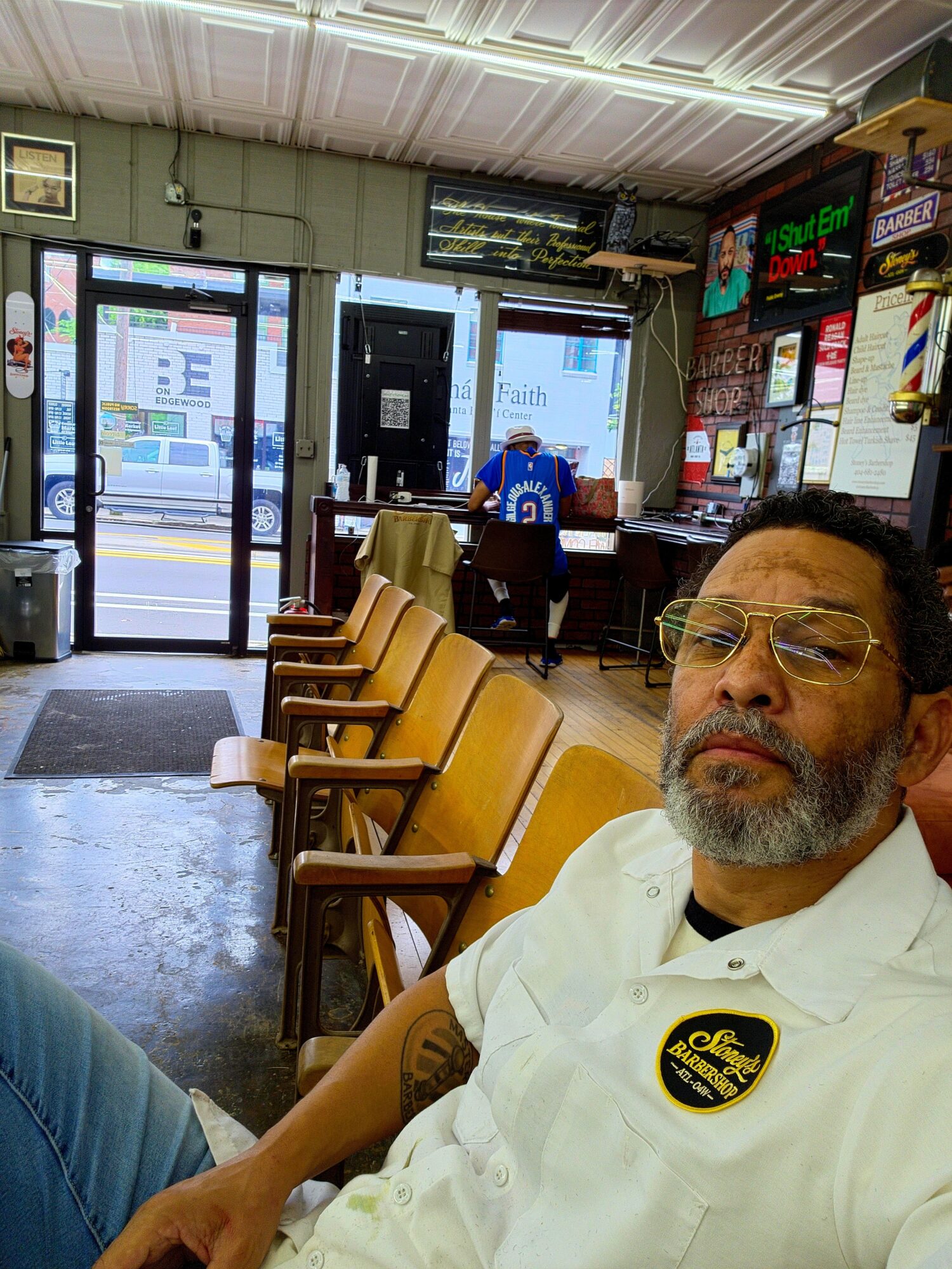 Man with glasses and beard sitting inside a restaurant, with chairs and a counter in the background, near a door and window.