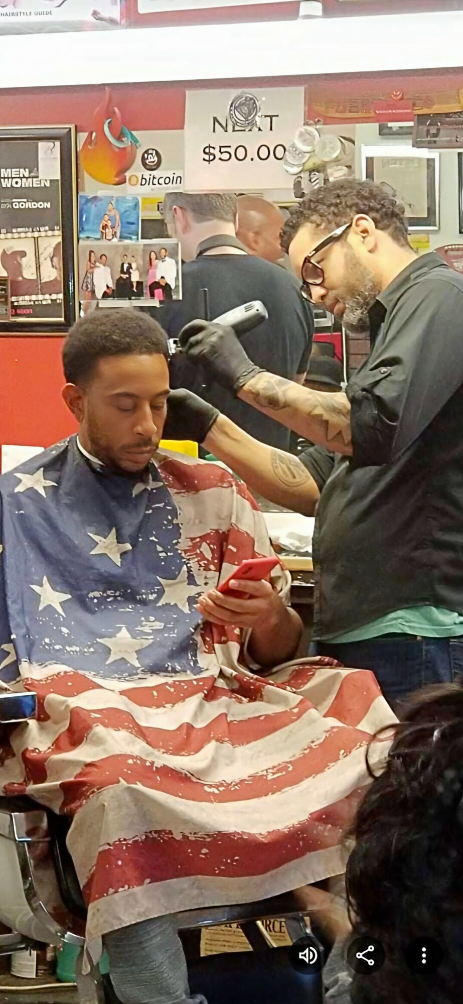 Barber cutting a man's hair, with an American flag cape, in a barbershop. Customers and signs visible in background.