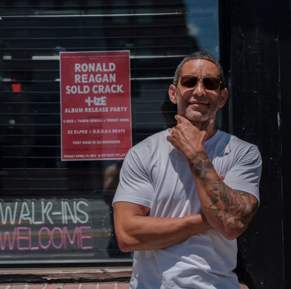 Man with sunglasses and tattoos smiling outside, with a red poster and neon sign behind him.