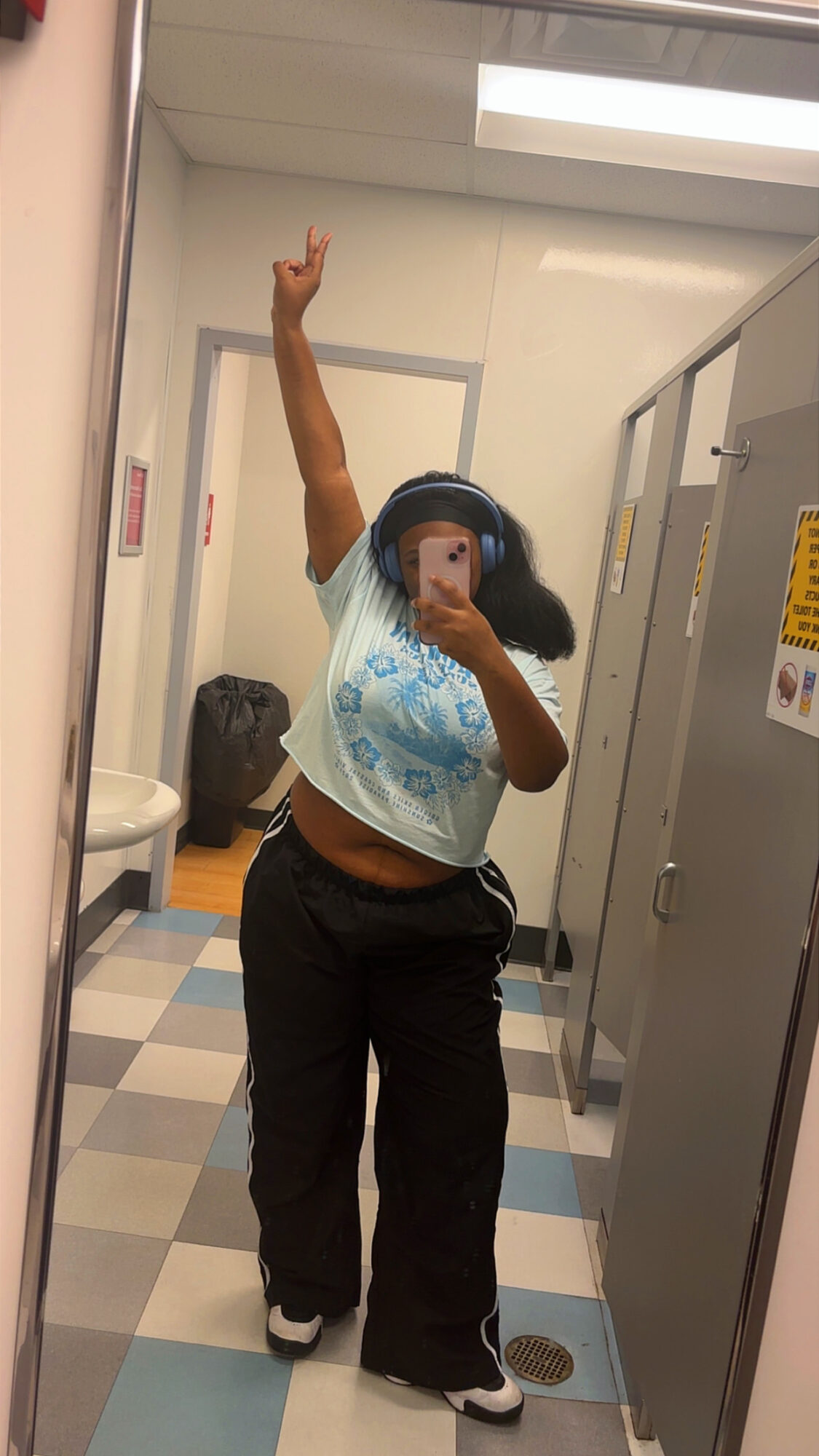Person taking a mirror selfie in a bathroom, raising one arm, wearing a light blue shirt and black pants.