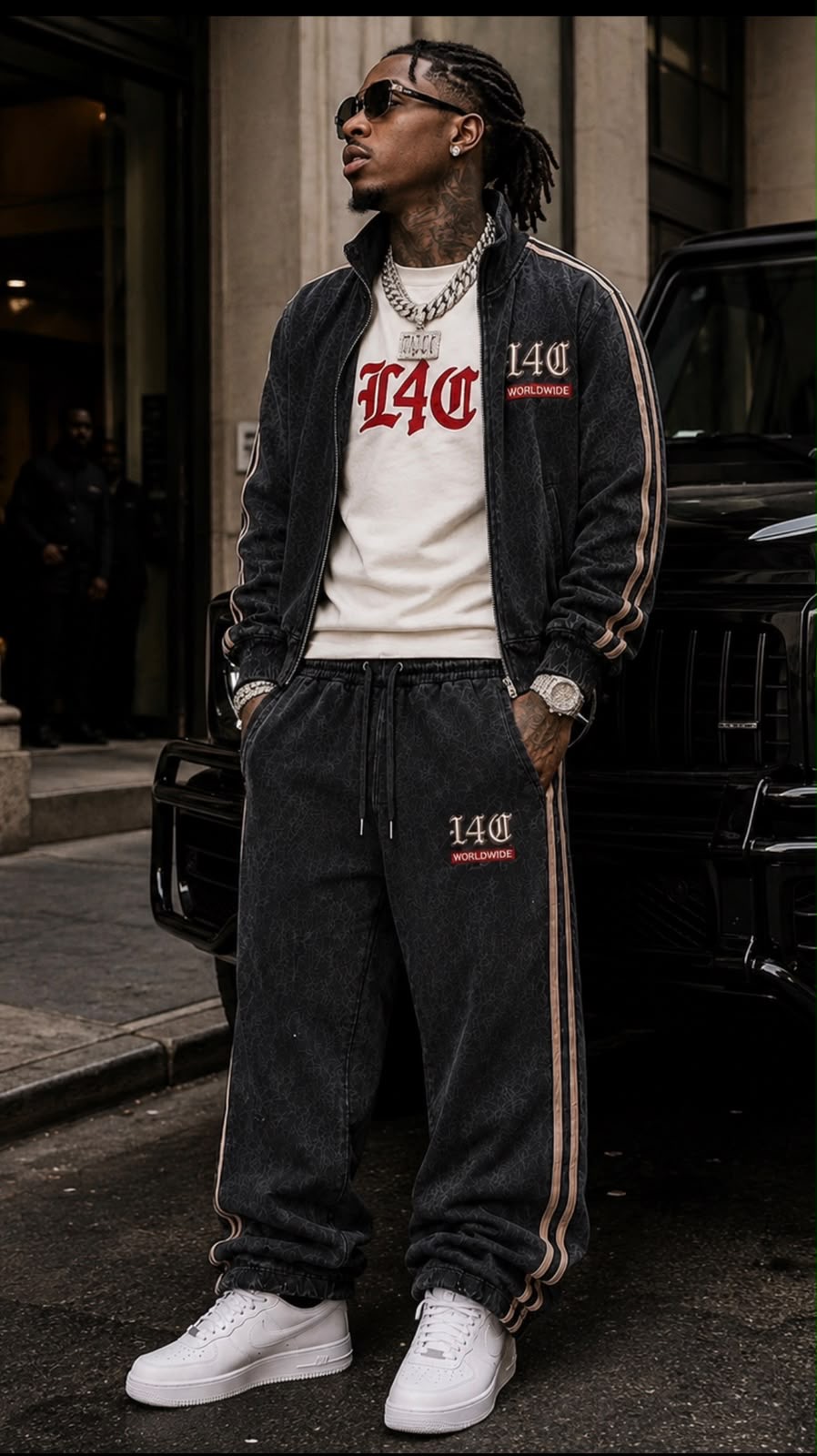 Man with sunglasses and jewelry stands on city street wearing black tracksuit and white sneakers.