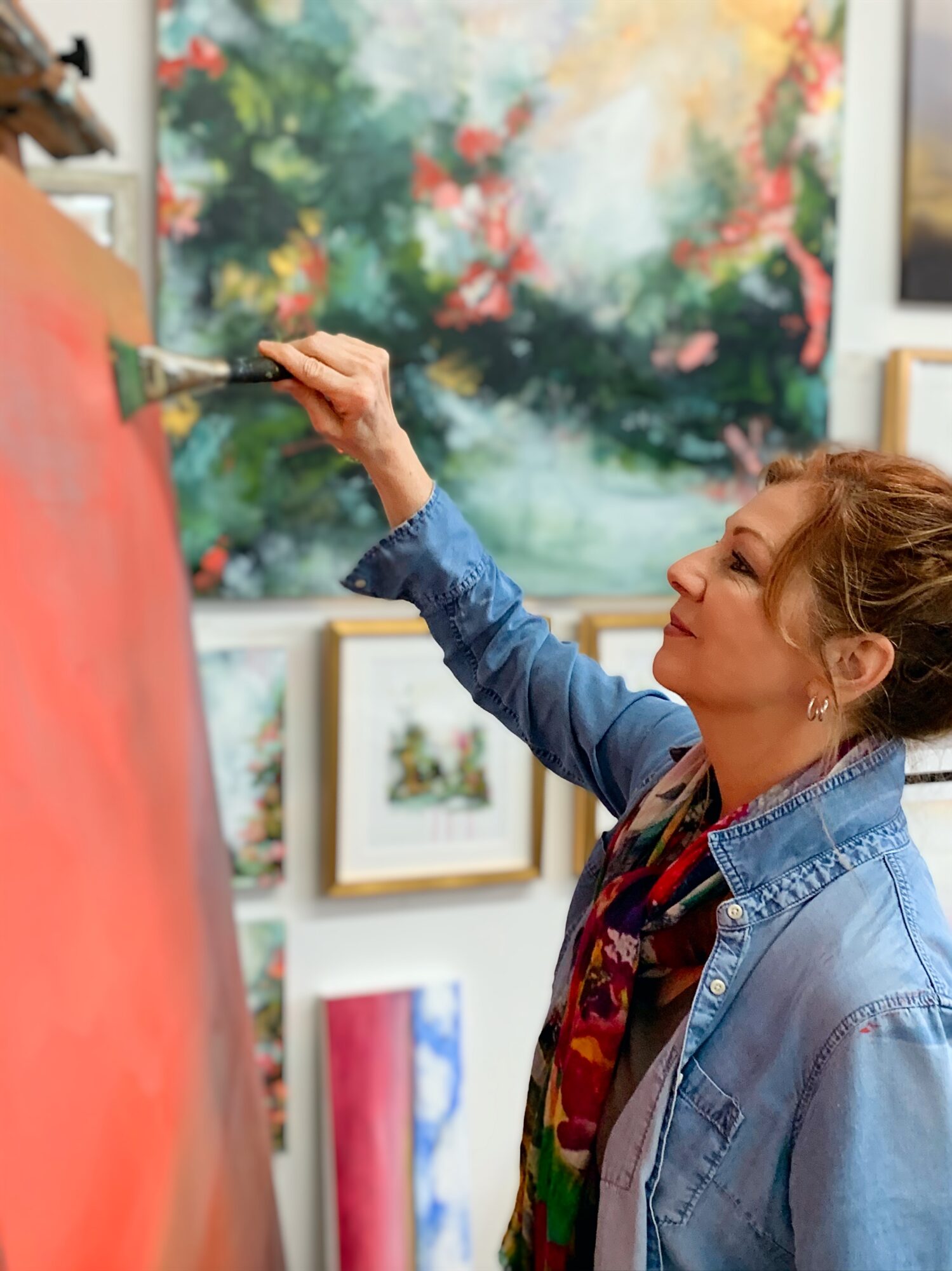 Woman painting on canvas in art gallery, colorful abstract artwork in background, wearing denim jacket, focused expression.