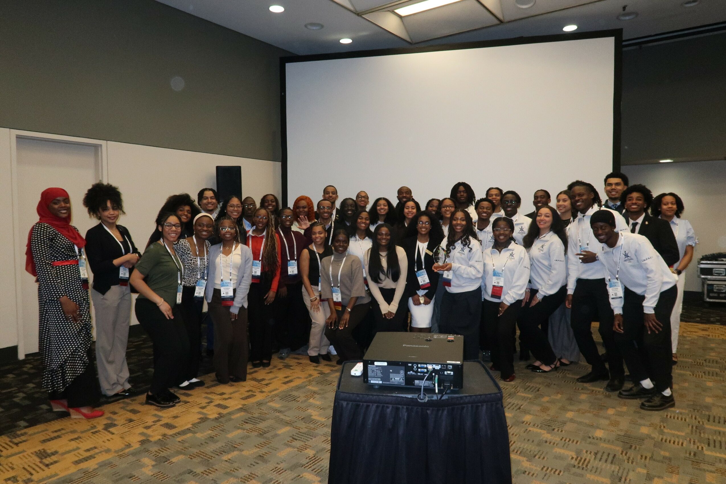 Group of people standing in a conference room in front of a large blank screen, some smiling and posing for the photo.
