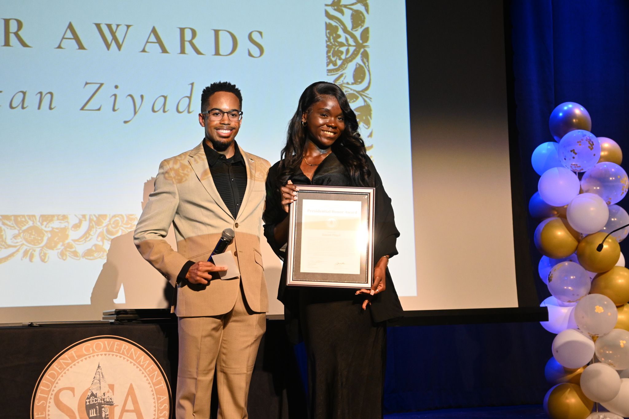 Two people on stage, one holding a framed award, smiling, with a large screen and balloon decorations in the background.