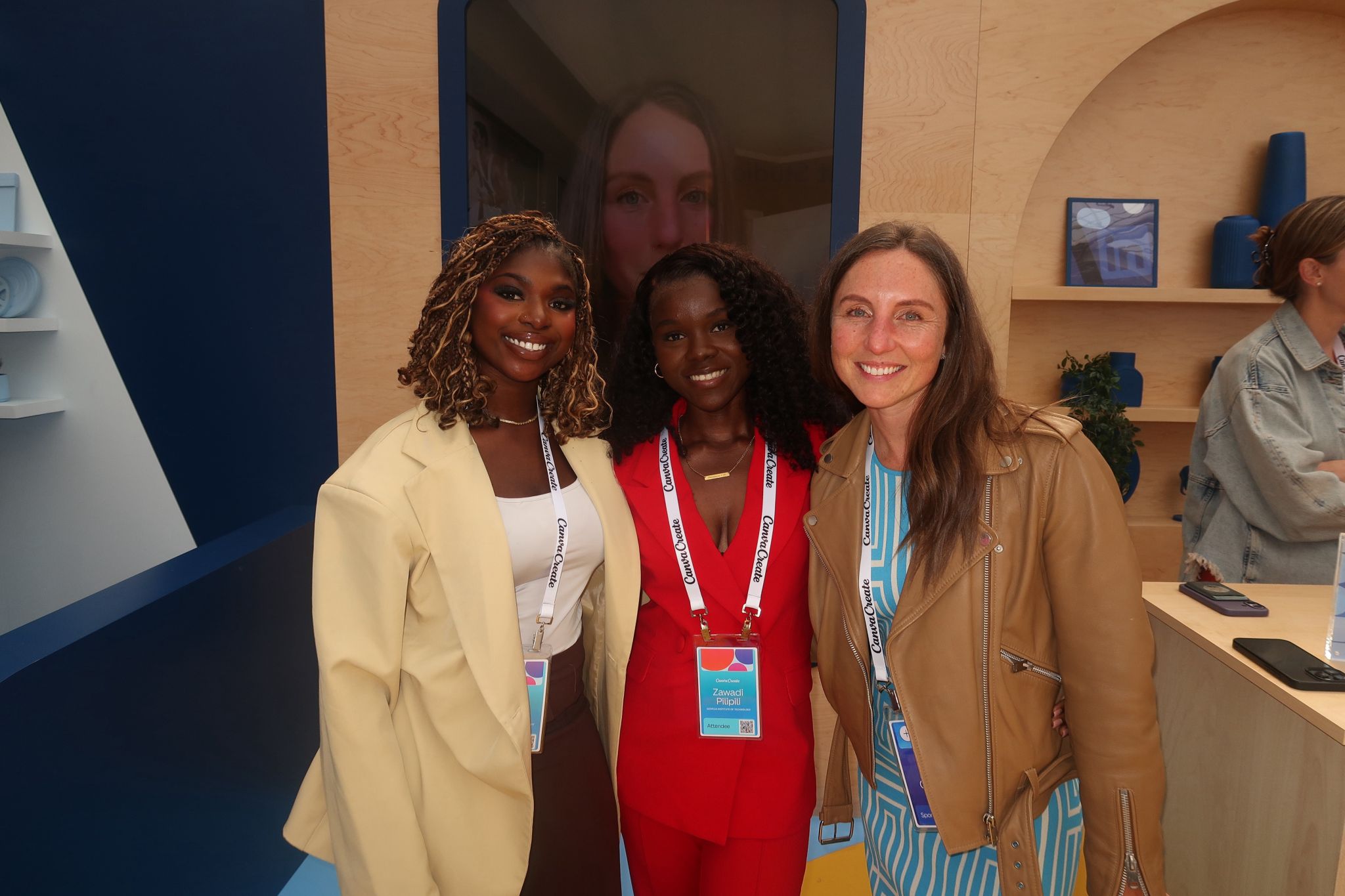 Three women smiling, standing together indoors with a large screen behind them, and a person in the background at a desk.