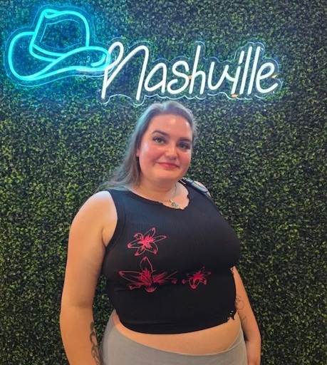 Woman standing in front of a green wall with neon Nashville sign, wearing a black sleeveless top with pink floral design.