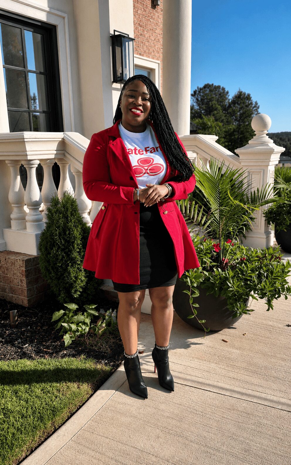 Young woman with long braids smiling outdoors, wearing a red coat, white graphic t-shirt, black shorts, and black boots, standing on a sidewalk with greenery and a building in the background.