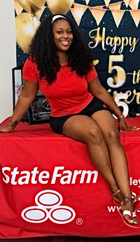 Woman in red shirt and black shorts sitting on a table at a birthday celebration, smiling, with a decorated backdrop.