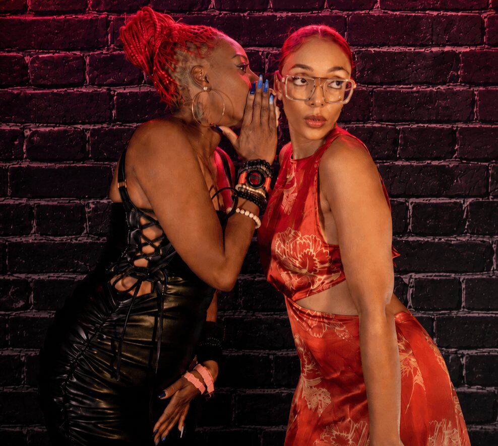 Two women with red hair and glasses stand against a dark brick wall, one whispering to the other, wearing stylish outfits.