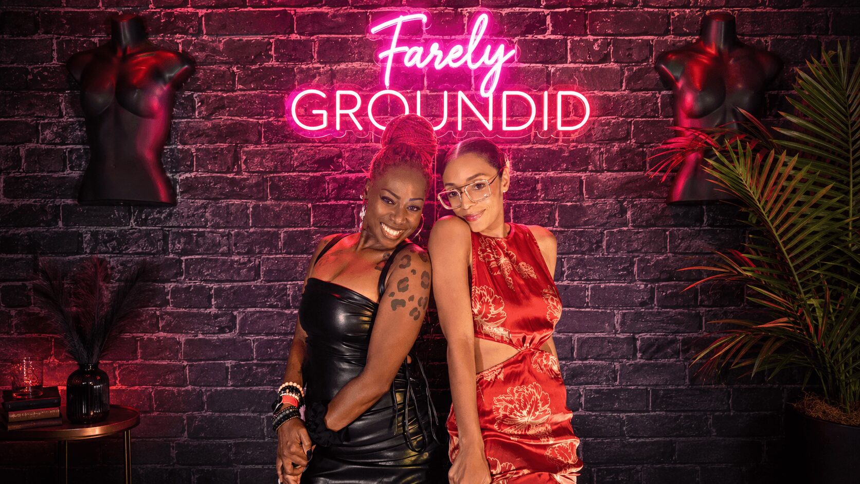 Two women pose in front of a brick wall with a neon sign that reads 'Farily Groundid,' surrounded by mannequins and plants.