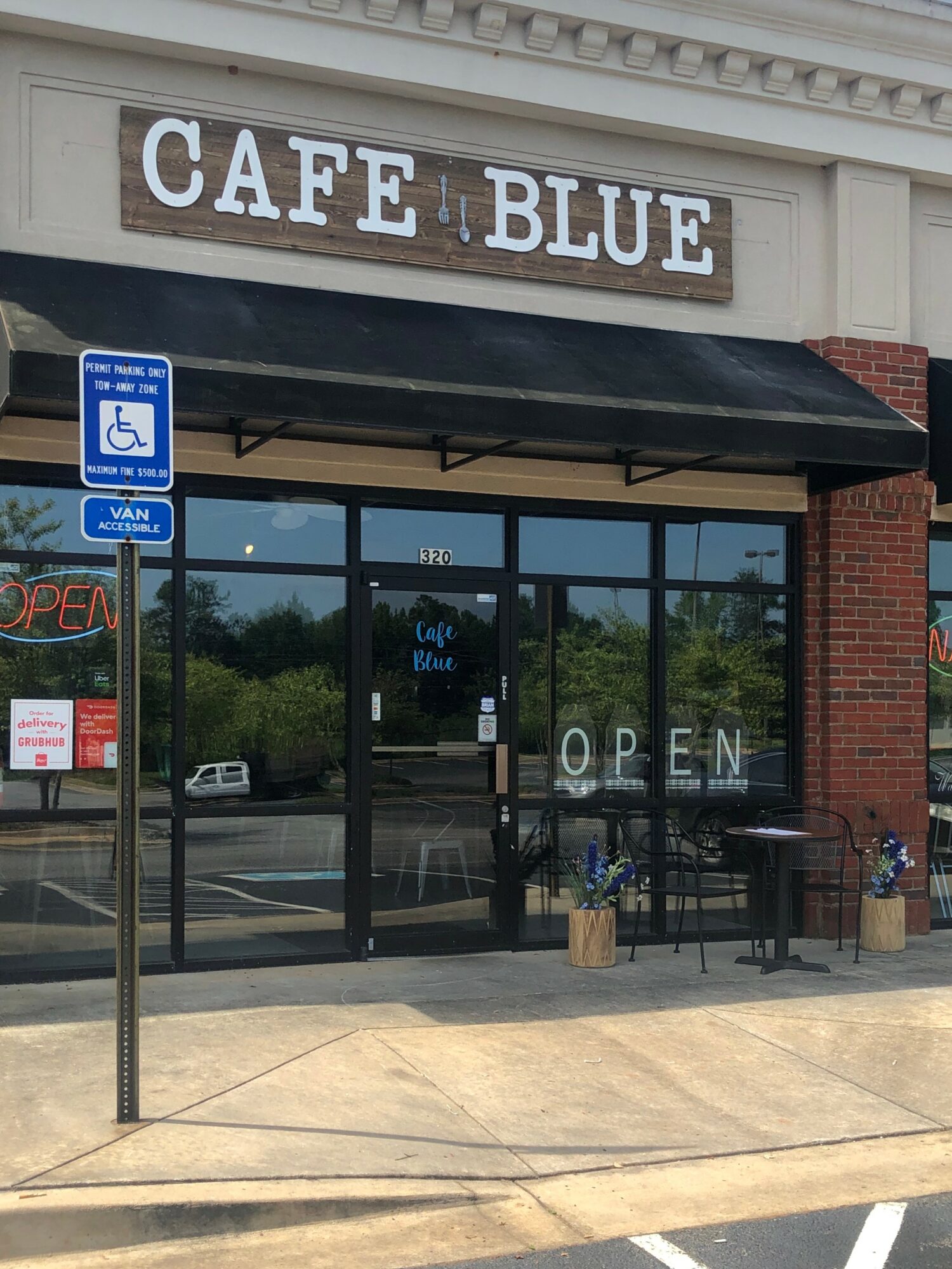 Cafe Blue storefront with glass doors, outdoor seating, and an 'Open' sign, reflecting trees and parking lot outside.