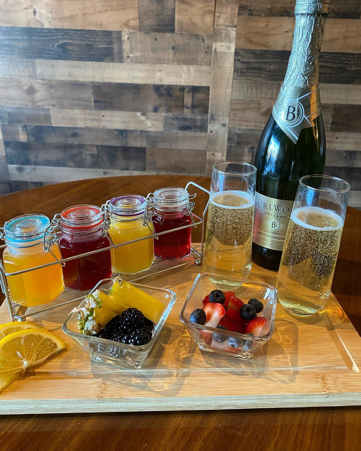 Assorted drinks and snacks on a wooden tray, including jars of colorful liquids, a bottle of sparkling wine, glasses, fruit, and berries.