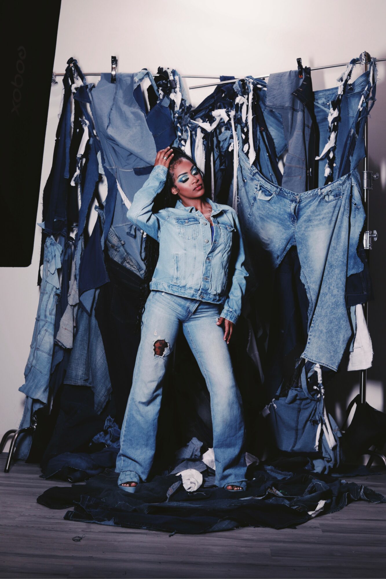 Person standing in front of a clothing display with jeans and denim jackets, holding their head with one hand, wearing sunglasses.