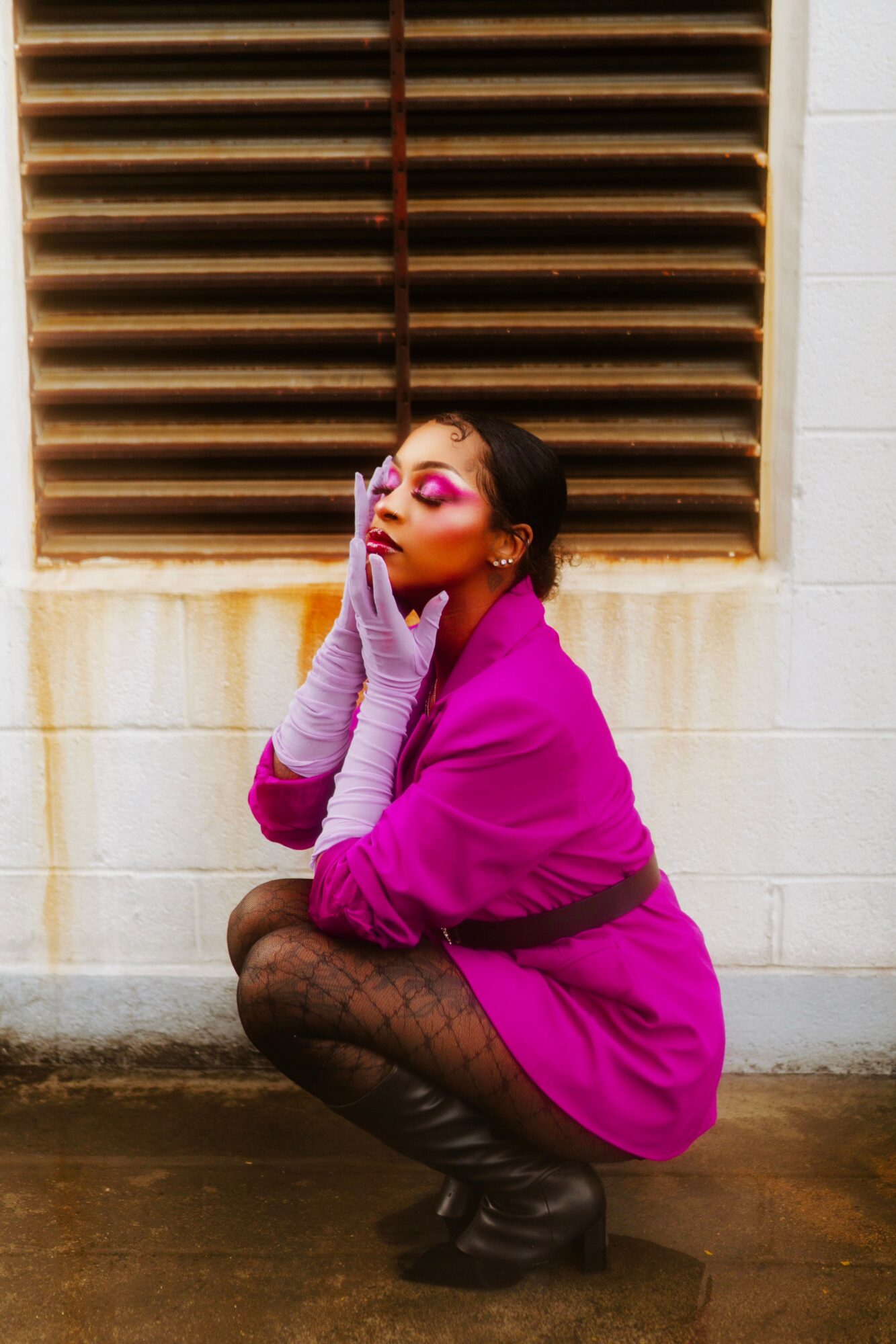 Person crouching with hands on face, wearing pink coat, patterned tights, and gloves, in front of a vented wall.