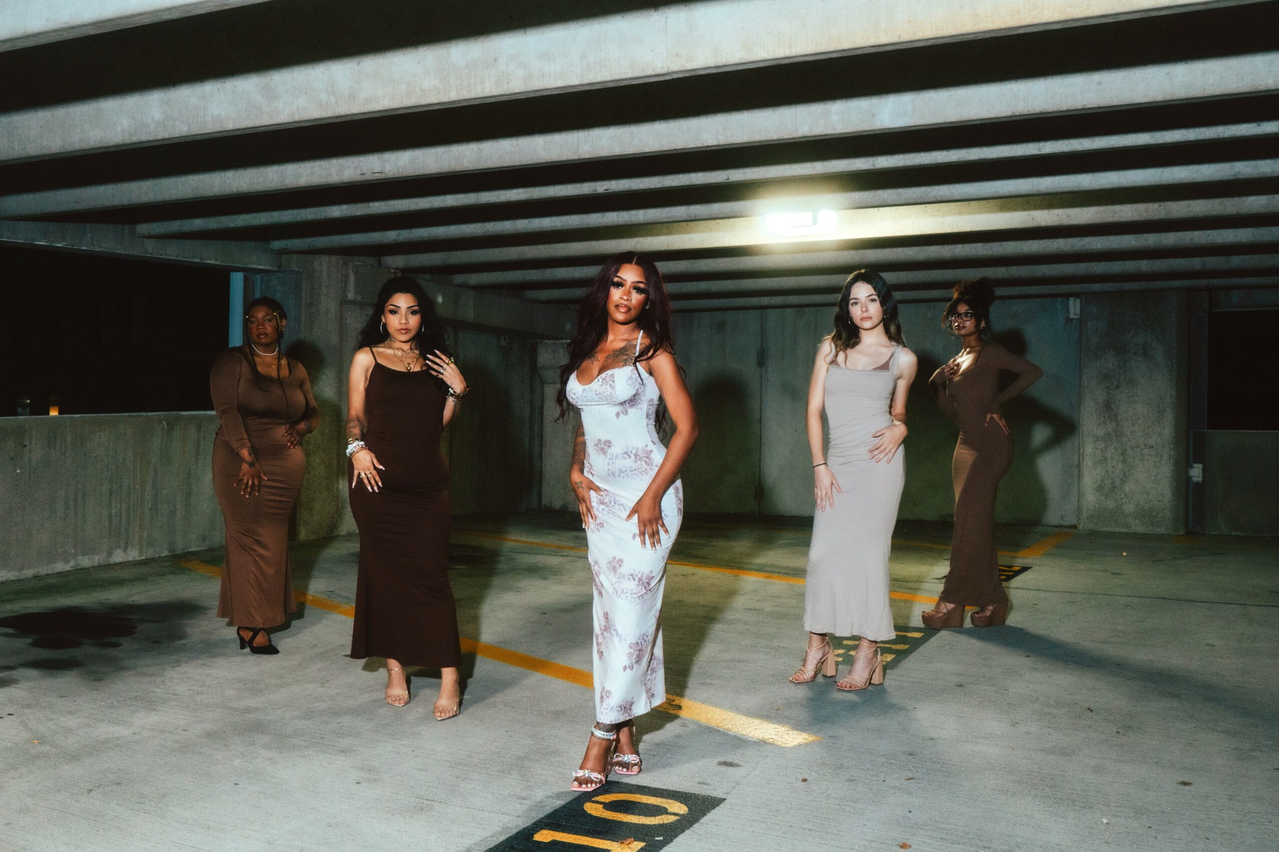 Five women in elegant dresses walk in a parking garage with concrete walls and ceiling, illuminated by overhead lights.