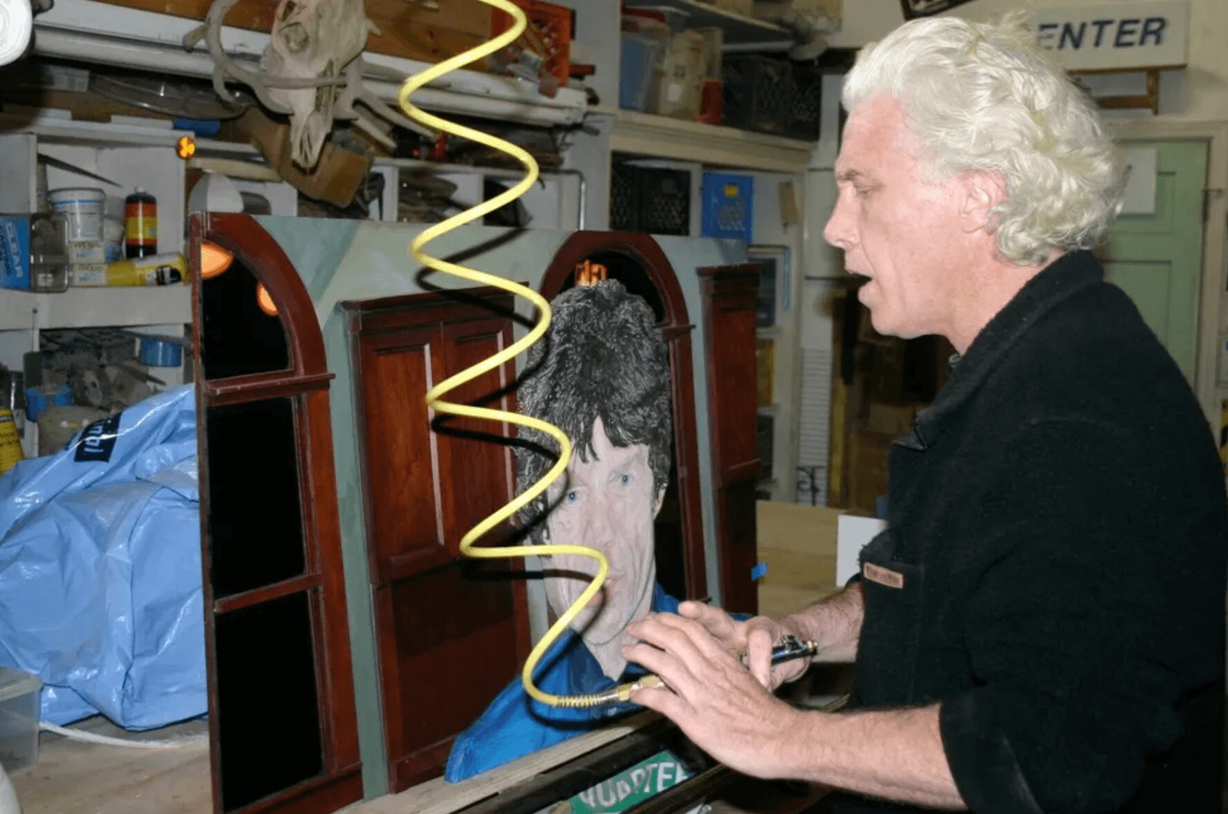 A woman with white hair looks at a mirror with a reflection of a face, in a cluttered workshop or storage room.