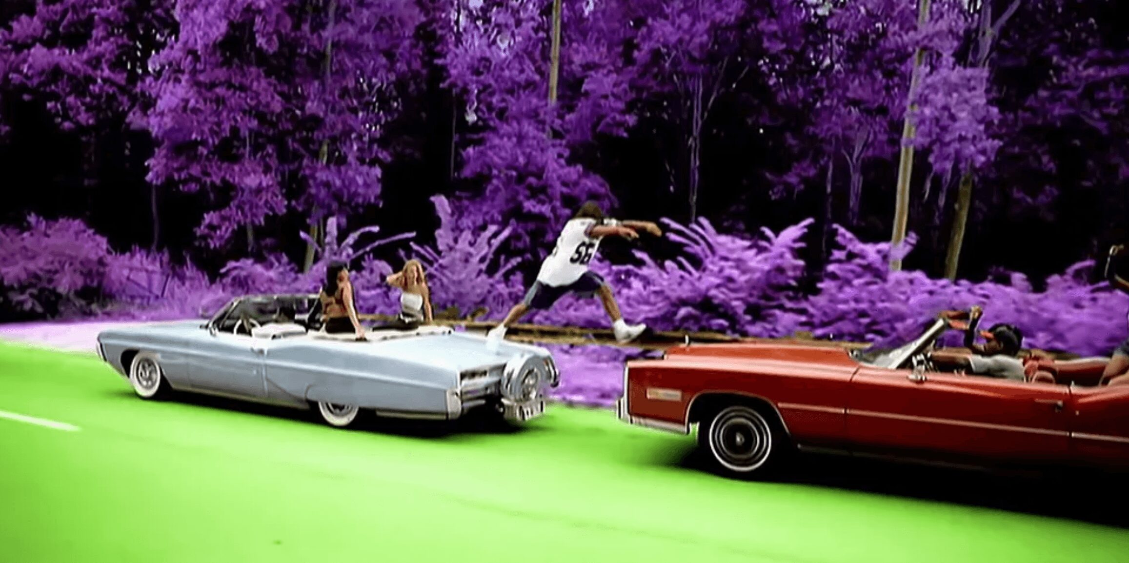 Two vintage cars on a road with purple trees in the background, one car is silver and the other is red, with people inside and around them.
