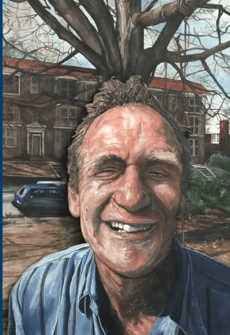 Smiling man outdoors with a large tree and buildings in the background.