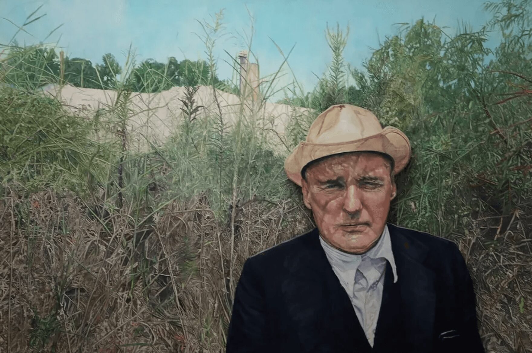 Man wearing a hat and dark jacket outdoors with tall grass and trees in the background.