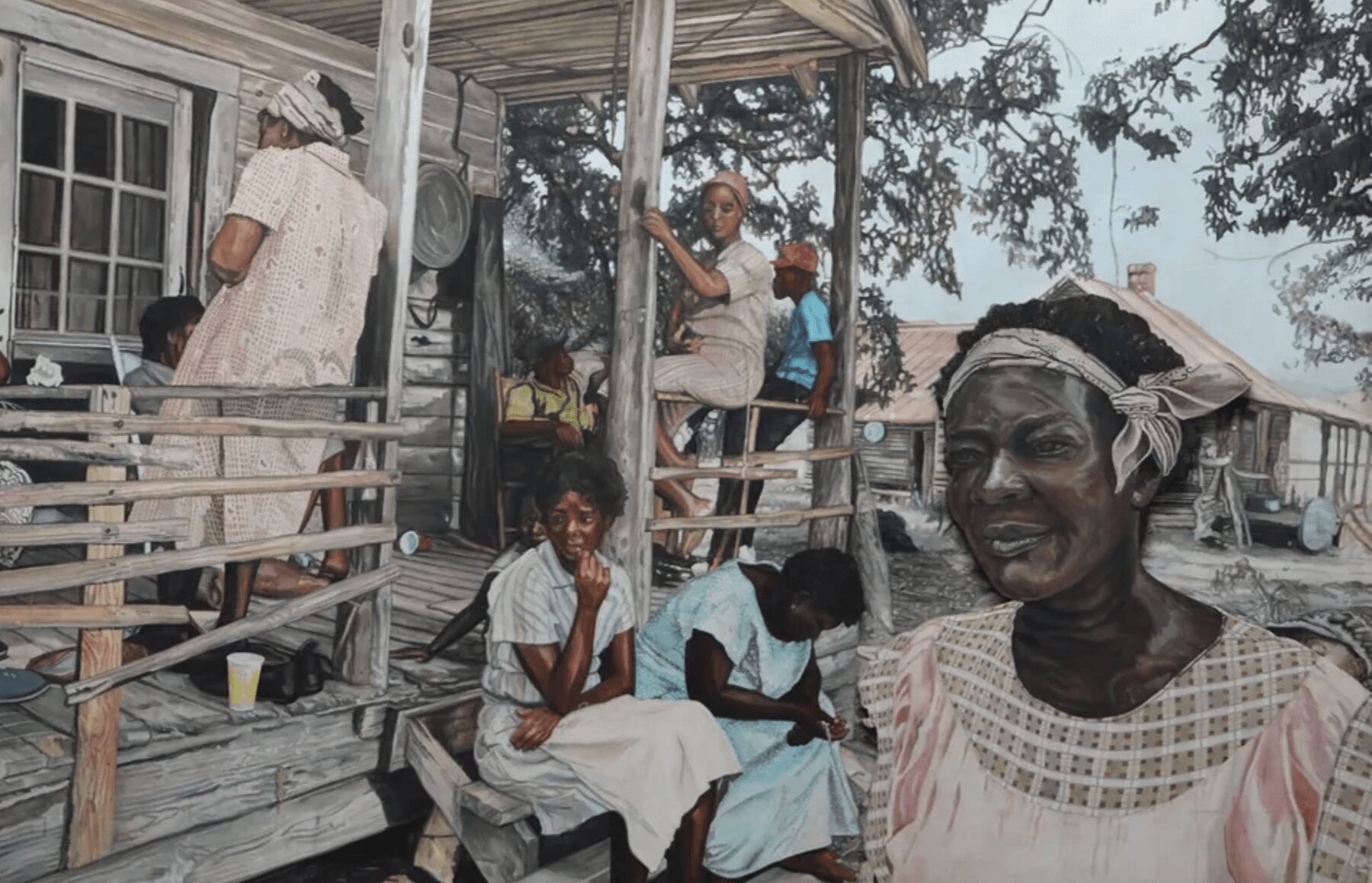 Group of people on a wooden porch, some sitting and some standing, with trees and houses in the background.