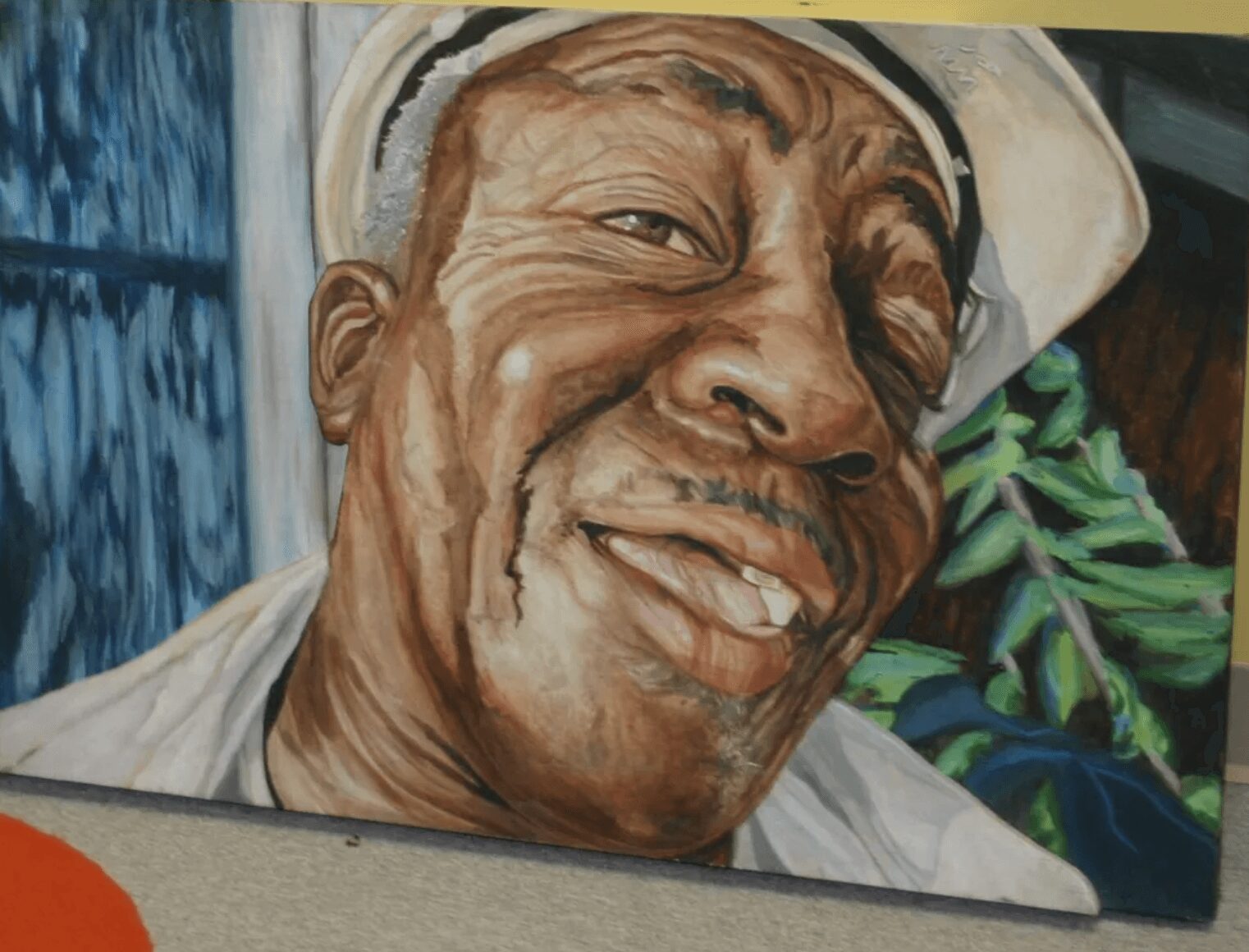 Close-up of a smiling elderly man wearing a white hat, with a background of green leaves and a blue curtain.