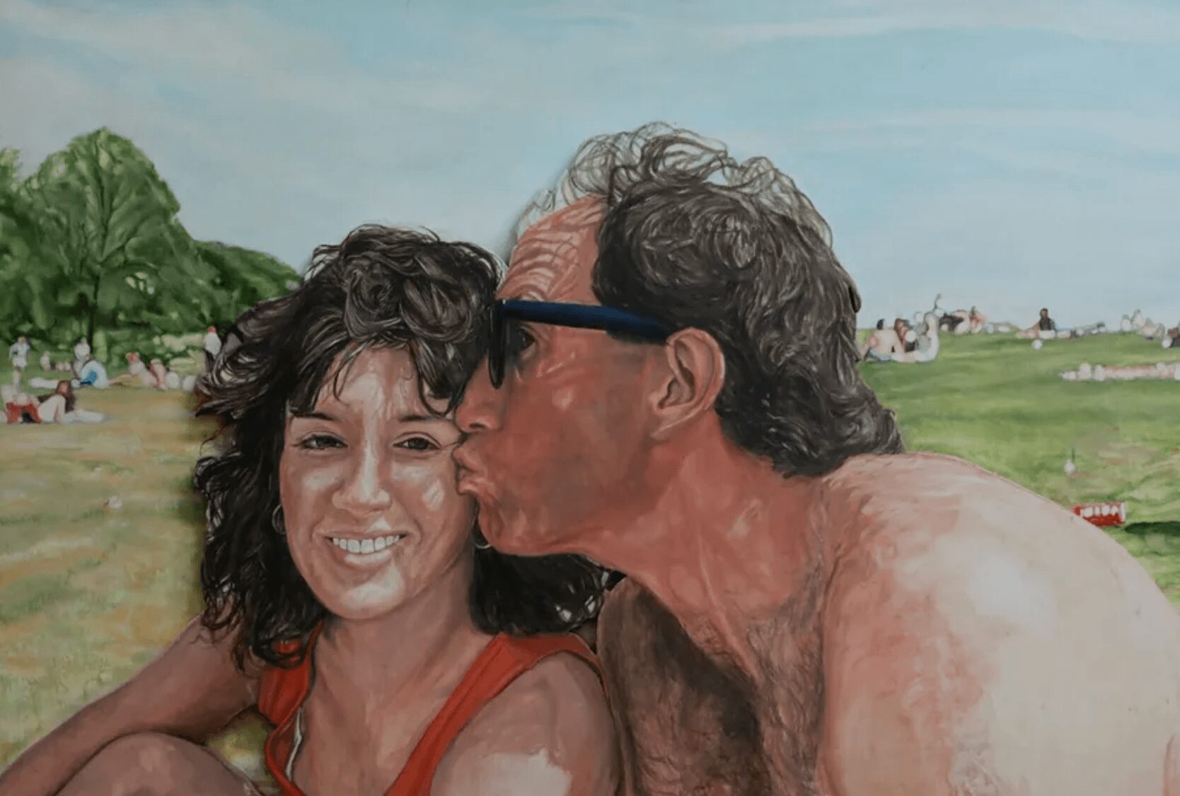 A man kisses a smiling woman on the cheek outdoors with a grassy field and trees in the background.